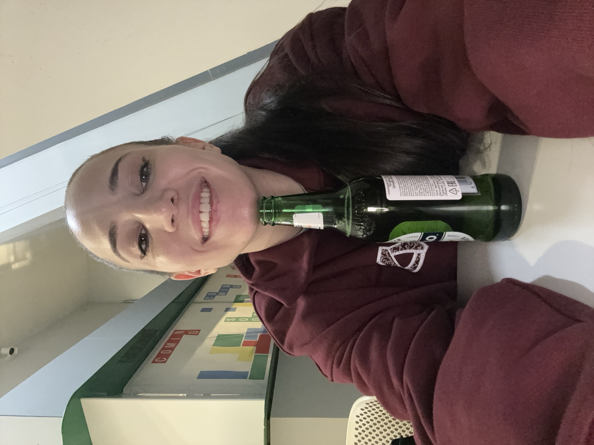 A woman with dark hair pulled back, wearing a maroon hooded sweatshirt, is depicted indoors in Moscow, Russia. She smiles directly at the camera. Her chin is positioned near the top of a green glass bottle, identified by its Cyrillic label as "Пиво светлое" (Light Beer) with a "ГОСТ" (GOST) standard mark and a volume of 0.33 liters. The bottle rests on a white table. In the background, a white wall features a green horizontal trim and a pattern of colored rectangular shapes (blue, yellow, green, red). Red, uppercase Cyrillic text "COMING SOON" is overlaid on this pattern. A white security camera is mounted on the ceiling in the upper left, and a white perforated chair back is partially visible to the woman's left.