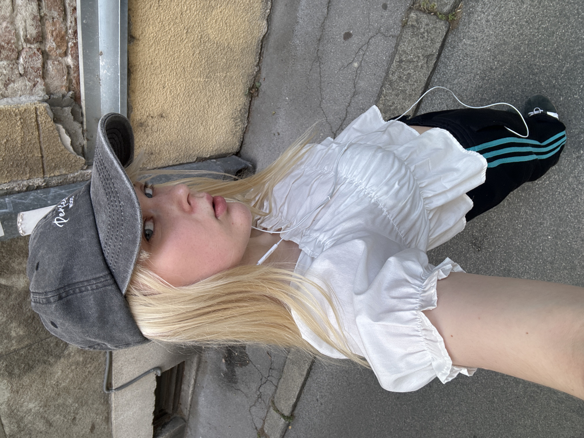 The image features a young person with blonde hair, wearing a grey baseball cap and a white ruffled blouse. They are also wearing black pants with three turquoise stripes down the side and white earbuds. The person is lying down on a paved surface, possibly a sidewalk, with a textured wall in the background. The scene appears to be outdoors, as suggested by the natural lighting and the presence of pavement and a brick wall. The overall impression is casual and relaxed, with the person looking upwards with a neutral expression. The presence of a white cord extending from the earbuds suggests they may be listening to music or a podcast. The time of day is not explicitly indicated, but the lighting suggests it is daytime. The location context is Vracar Urban Municipality, Serbia. No specific event or activity is clearly depicted, other than the person resting. The text visible on the cap is partially obscured but appears to be a logo or brand name.