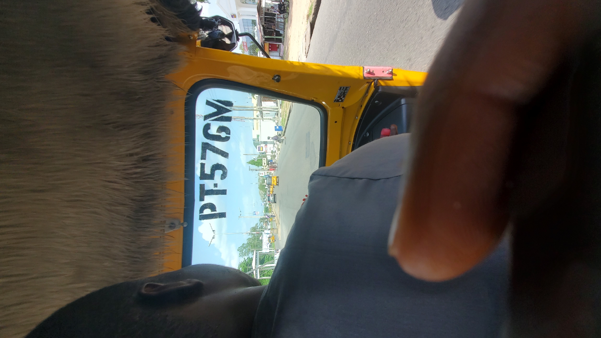 The media file is a first-person perspective shot taken from inside a yellow auto-rickshaw in Jalingo, Nigeria. The main subjects are the interior of the auto-rickshaw, the driver's shoulder and head (partially visible), and the road ahead seen through the side window. The driver's hand or arm is prominently in the foreground, blurred and out of focus. The scene depicts a street in what appears to be a semi-urban or town setting, with buildings lining the road, other vehicles, and pedestrians visible in the distance. The weather seems to be clear and sunny, suggesting it is daytime.
The activity taking place is travel by auto-rickshaw. The perspective suggests the auto-rickshaw is moving, possibly on a road with moderate traffic. Notable details include a furry, light-brown material on the left side of the frame, which might be part of the auto-rickshaw's interior or a seat cover. On the side window, the text "PT576GM" is clearly visible in large, black capital letters. The overall mood is candid and informal, capturing a moment of everyday travel. The location context of Jalingo, Nigeria, is provided, which helps in understanding the type of vehicle and the environment.