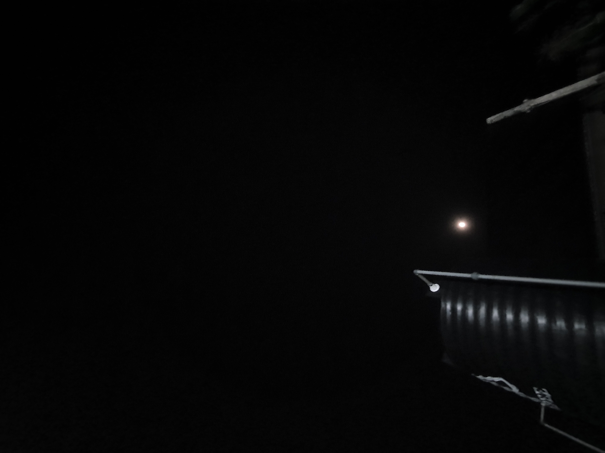An outdoor, low-light, close-up, angled shot shows the top of a large, dark, ribbed water tank with a light fixture attached to a pole extending from the tank. The light fixture is illuminated. A faint, circular object, possibly the moon, is visible in the dark sky to the right of the tank. The bottom right of the frame shows a blurry, vertical wooden pole. The scene is dark, indicating it is nighttime.