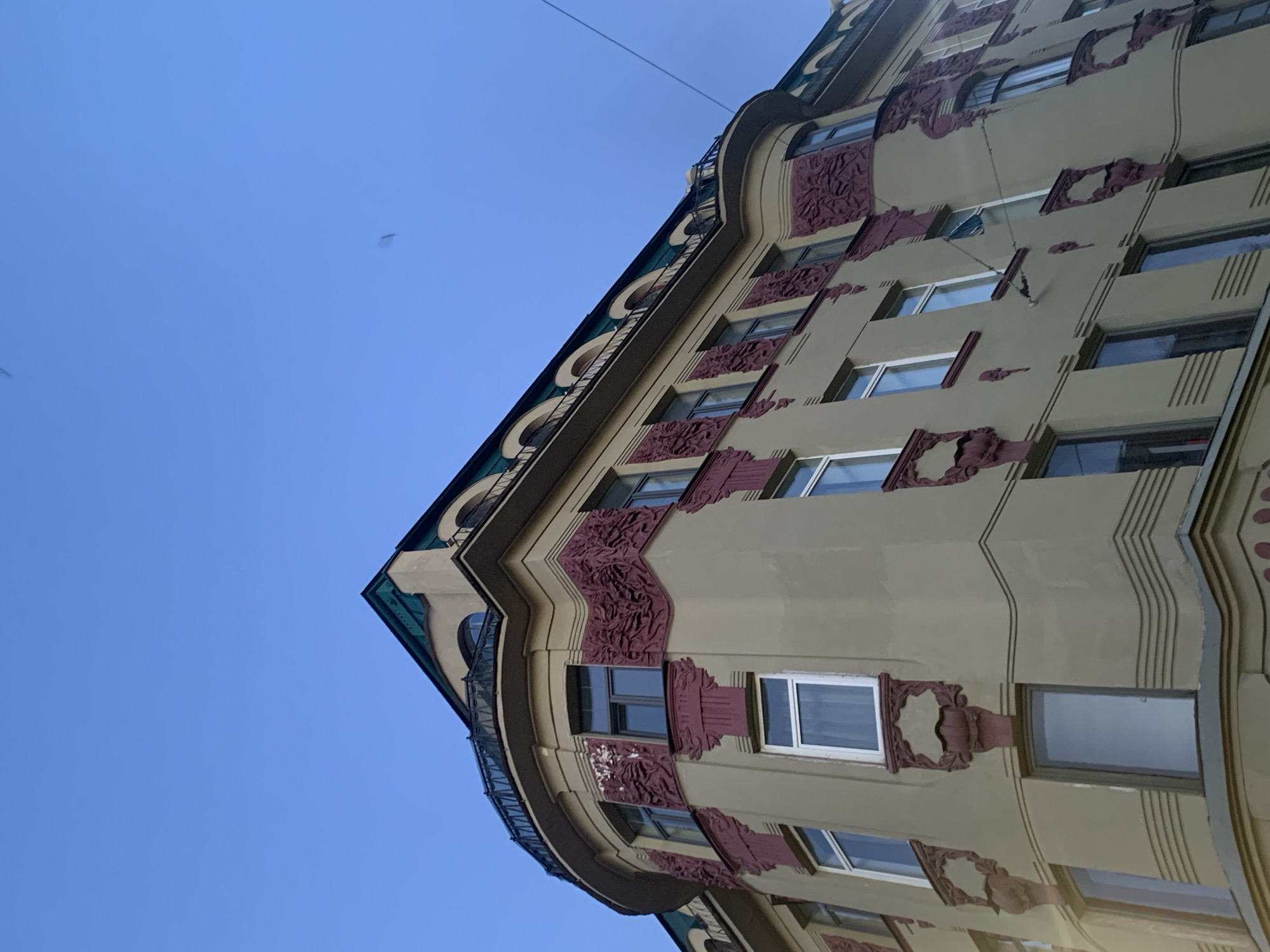 This is a low-angle, full shot of the corner of a tan-colored building with ornate burgundy architectural details, set against a clear blue sky. The building features multiple windows, some with white frames and others with dark frames. The architectural style is reminiscent of Art Nouveau. A rounded balcony with a dark metal railing is visible on the upper floor. The roofline is dark green with arched windows. The building appears to be located in Saint Petersburg, Russia, likely within the historic city center given its architectural style. There are no people or vehicles visible in the frame. A thin wire runs across the upper right portion of the image.