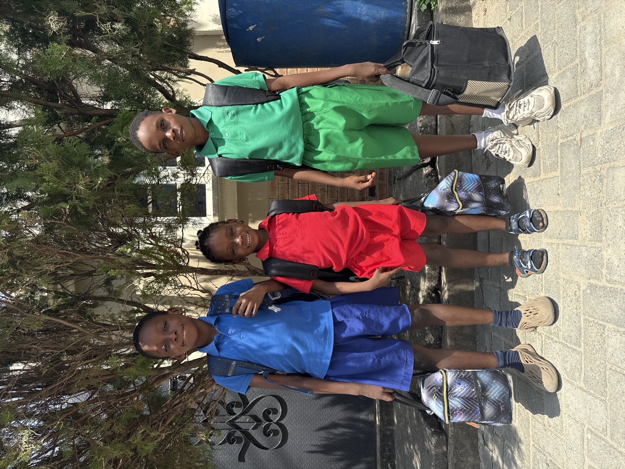 Three children in school uniforms stand in a row outside, with a metal gate and part of a building visible behind them. The child on the left wears a blue shirt and shorts, the one in the middle a red dress, and the child on the right a green shirt and shorts. Each child carries a backpack. The setting appears to be a residential area with trees and paved ground. The city is Ado, Nigeria.