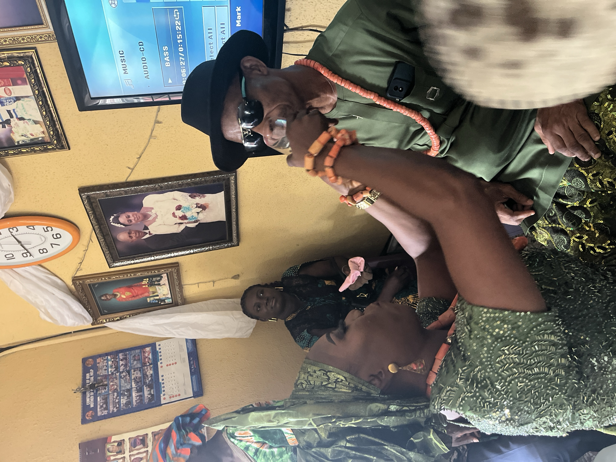 The image captures an indoor scene likely in a home in Warri, Nigeria. A man wearing a green shirt, black hat, and sunglasses, along with an orange coral bead necklace, is interacting with a woman adorned with similar orange bead jewelry, and wearing an olive-green dress. Another woman is seated behind them. A TV screen displays music options. The walls are decorated with framed photographs, a clock, and a calendar bearing the title "Confraternity of Our Mother of Perpetual Help".