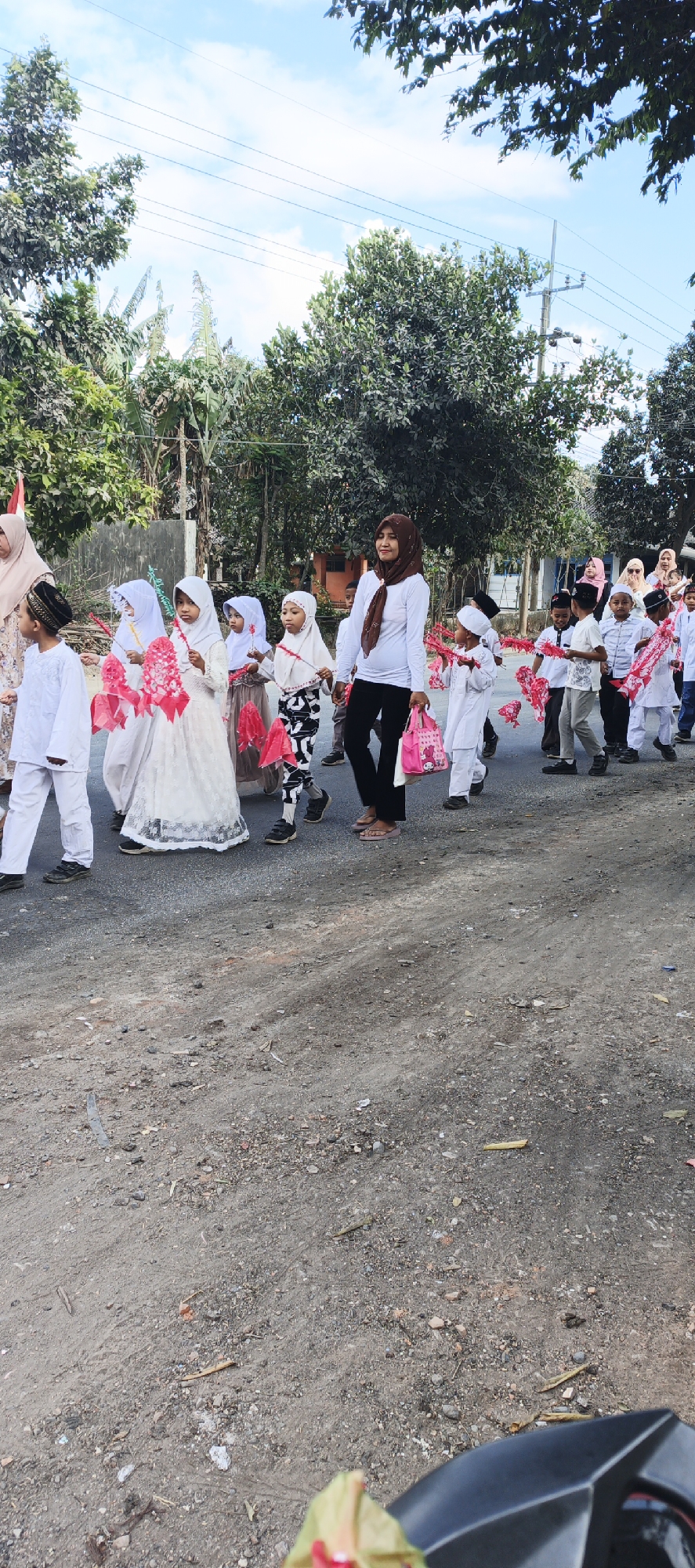 This image shows a procession of children and adults walking down a dirt road in Grenden, Indonesia. The children are dressed in white Islamic attire and some are carrying decorative pink paper flowers. A woman in a white long-sleeved shirt and brown headscarf is walking alongside them, holding a pink bag. Trees line the road, and buildings are visible in the background. The sky is blue with some clouds. The ground is rough and uneven, with small stones and debris.
