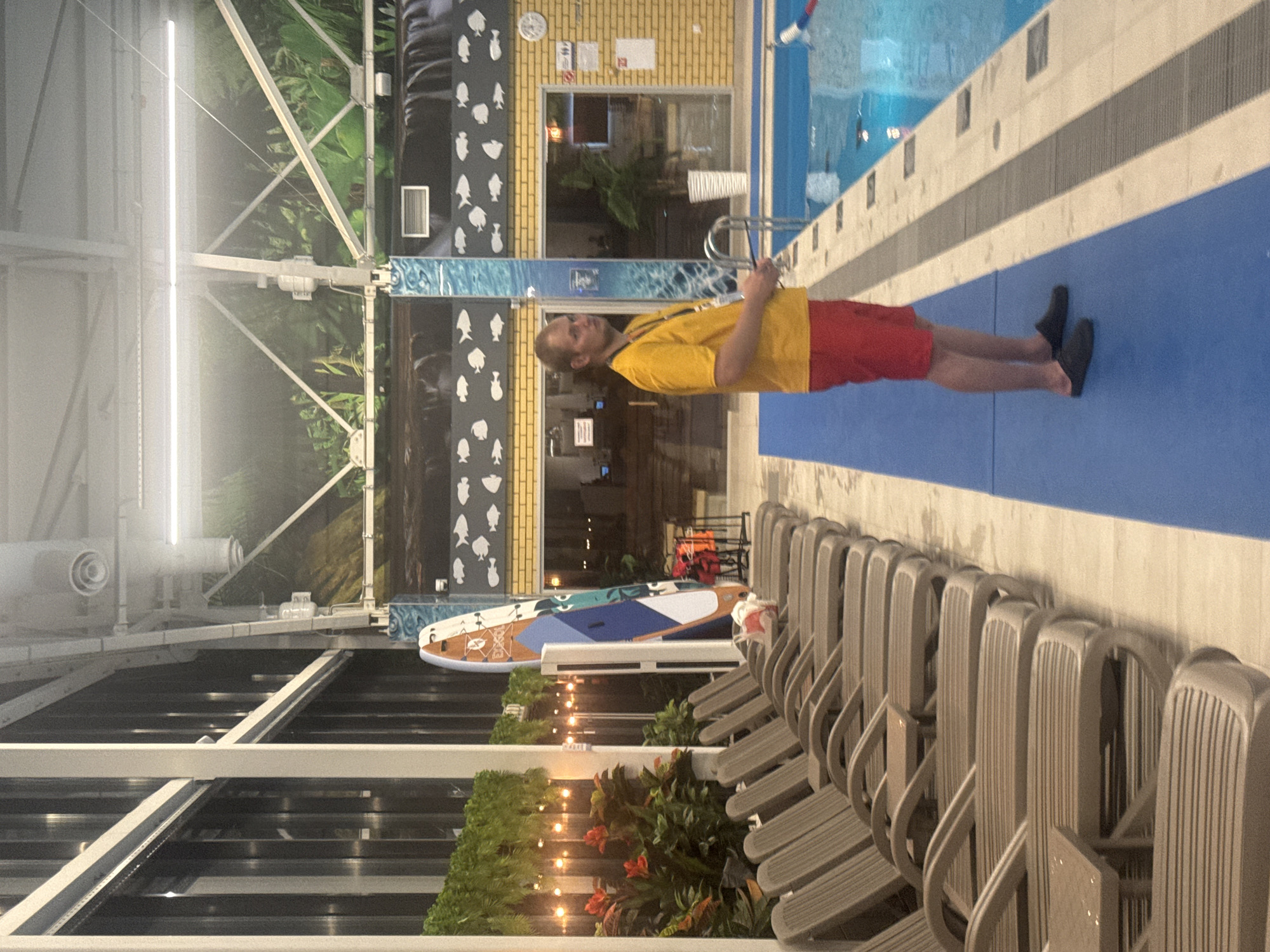 A lifeguard stands watch by a pool indoors. The scene features rows of empty lounge chairs and decorations of plants. In the background there is a window and a wall decoration featuring spade-shaped icons. The location is potentially a water park or pool in Ulyanovsk, Russia, as the lifeguards wear the traditional colors yellow and red.