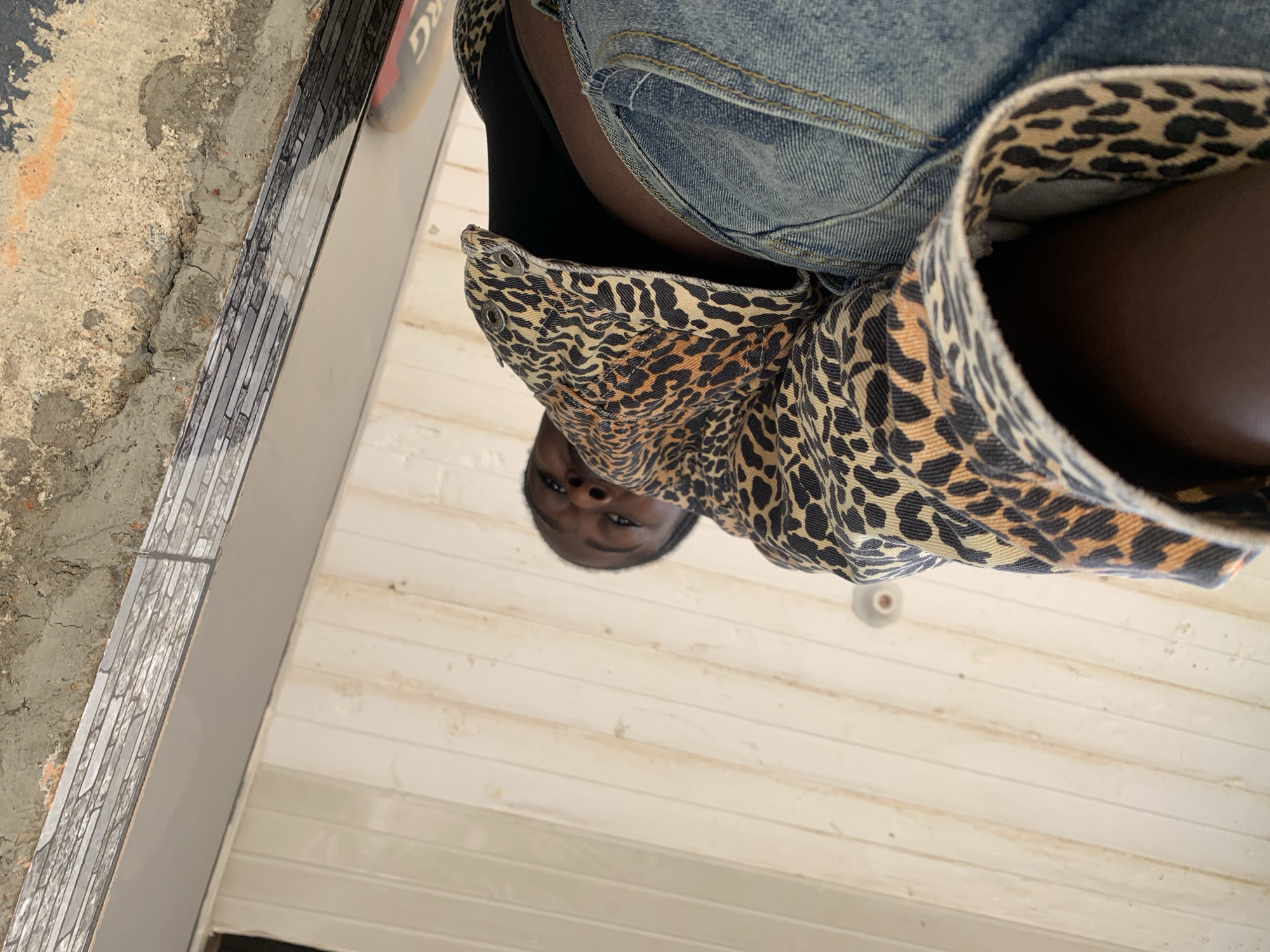 This image, captured in New Stadium, Nigeria, presents an individual from a low-angle perspective, looking upwards. The subject, a person with dark skin, is visible from the upper torso, wearing a leopard print jacket over a black undergarment and blue denim jeans. Their gaze is directed towards the camera. The background consists of a light-colored, vertically paneled surface that occupies the lower and left portions of the frame. Above this, a rough, unfinished concrete or cement structure is visible, characterized by cracks and varied texture. A horizontal trim piece with a grey, brick-like pattern separates the paneled surface from the concrete. A small, circular, light-colored fixture is affixed to the paneled surface. In the far upper right, a dark object with partially visible white lettering (possibly "DKG") is faintly discernible.