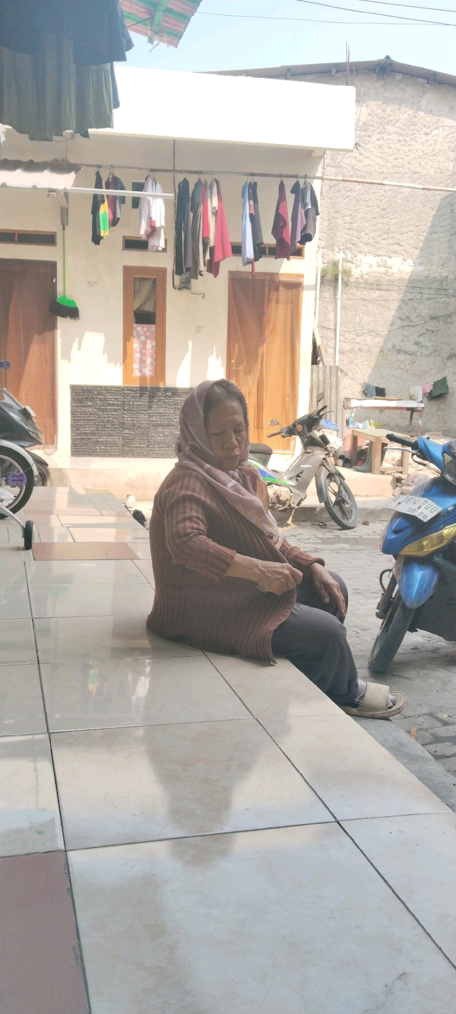 The image shows an elderly woman wearing a headscarf and a brown cardigan sitting on tiled steps outside a building. Clothes are hanging on a clothesline in the background, and two motorcycles are parked nearby. The scene appears to be a residential area in West Jakarta, Indonesia. The woman is looking down and appears to be adjusting her clothing. The environment is sunny, with shadows cast on the ground.