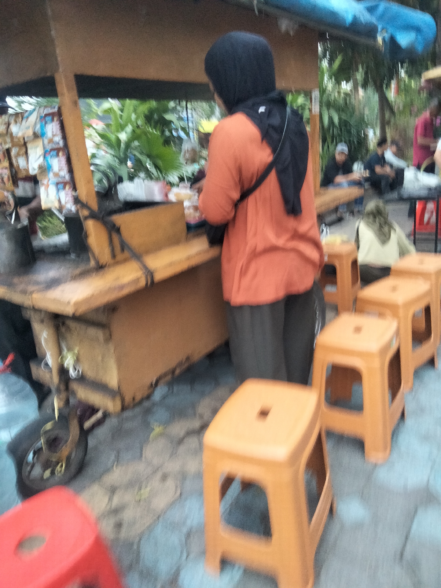 The image depicts an outdoor food stall in Surakarta, Indonesia, with a focus on a woman wearing a black hijab and an orange shirt. She is standing behind a wooden food cart, which has various packaged goods displayed on its side. Several orange plastic stools are arranged in front of the cart, indicating a seating area for customers. In the background, other individuals are visible, some seated at tables and others standing. Lush green plants and trees form a natural backdrop to the scene. The ground is paved with interlocking stones.