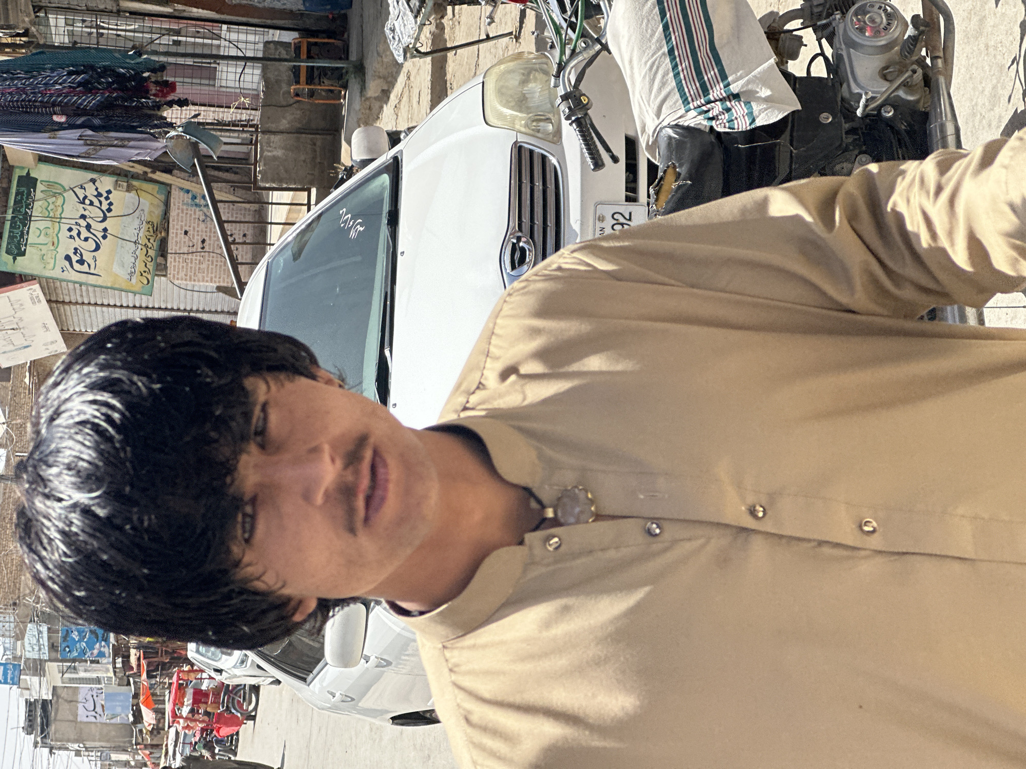 In Kanozai, Pakistan, a young man with dark, tousled hair and a thin mustache stands in the foreground, looking directly at the camera. He is wearing a light brown collared shirt with two visible buttons at the neck, and a necklace with a round, ornate pendant. Behind him, a white car is parked on a dusty street lined with shops and buildings. The street is bustling with activity, with other vehicles, including a motorcycle partially visible to the right, and various awnings and signs in the background, suggesting a market or commercial area. The overall impression is a candid street portrait taken on a bright, sunny day.