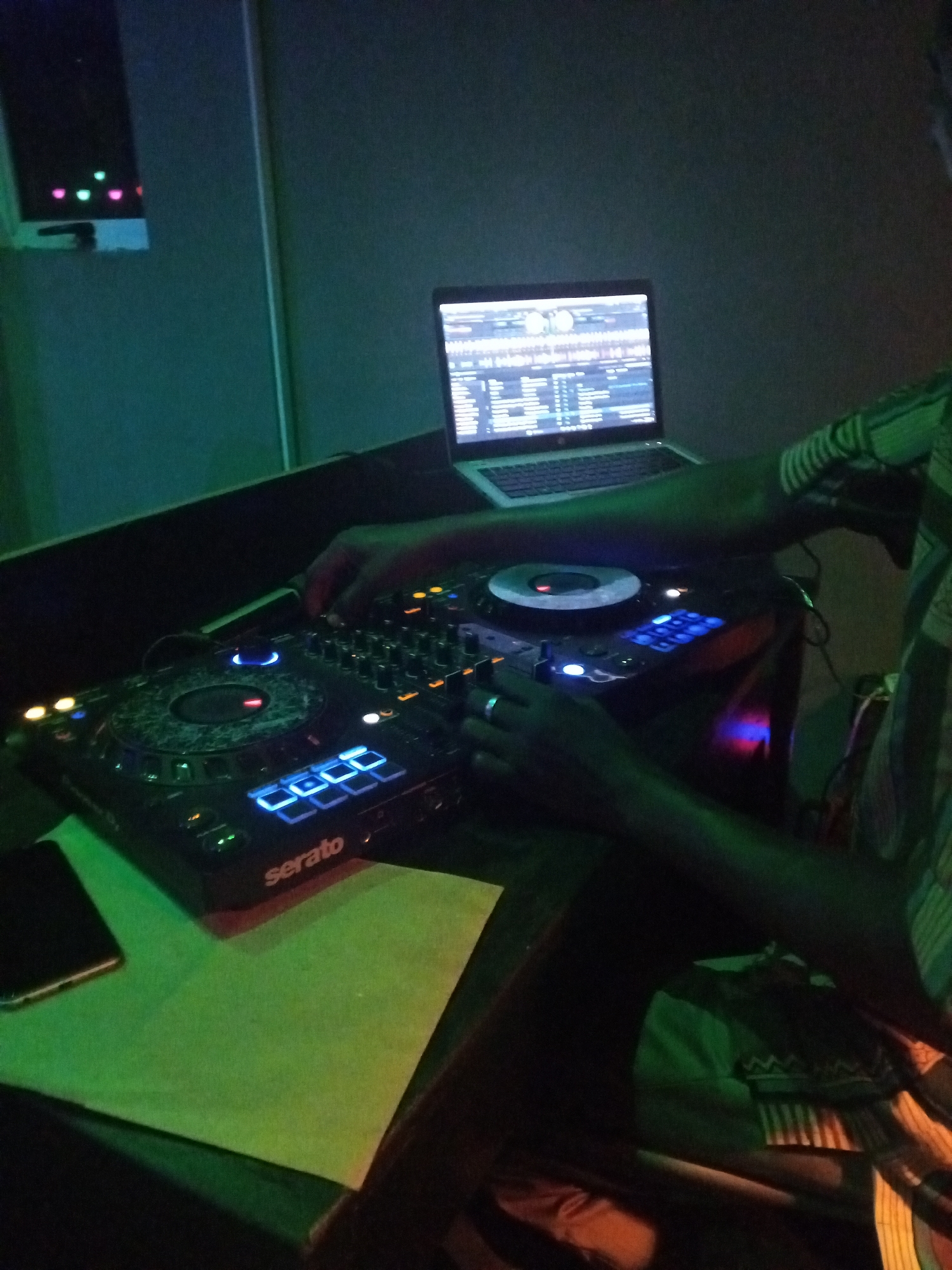 A DJ is working on a Serato DJ controller with a laptop showing DJ software. The location appears to be a dark club or studio with a monitor reflecting green and pink lights on the wall. A phone and a piece of paper are on the table next to the DJ controller. The DJ is wearing a patterned shirt. It is not possible to confirm that the location is in Lokoja, Nigeria.