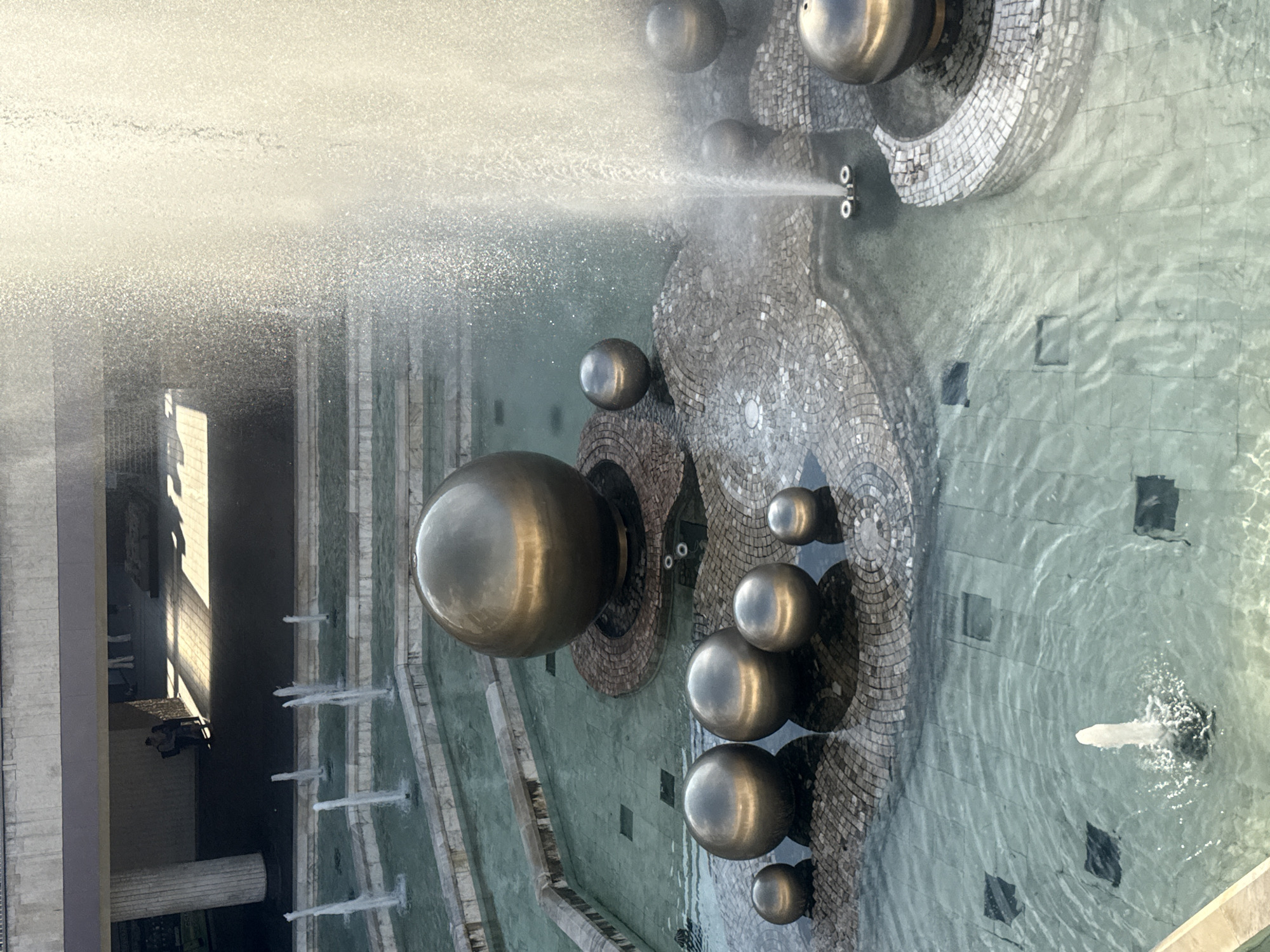 The image depicts an outdoor fountain with multiple tiers and several metallic spheres of varying sizes. In the foreground, a large metallic sphere sits prominently, surrounded by smaller spheres arranged on a mosaic-tiled platform. Water jets are visible, spraying upwards and creating a misty effect. The fountain is situated in a tiered pool with clear, shallow water. In the background, two indistinct figures are visible under an overhang, suggesting the presence of an urban environment. The architectural style hints at a public space in Sofia, Bulgaria, likely a square or park. The overall impression is one of a modern public art installation integrated with water features.