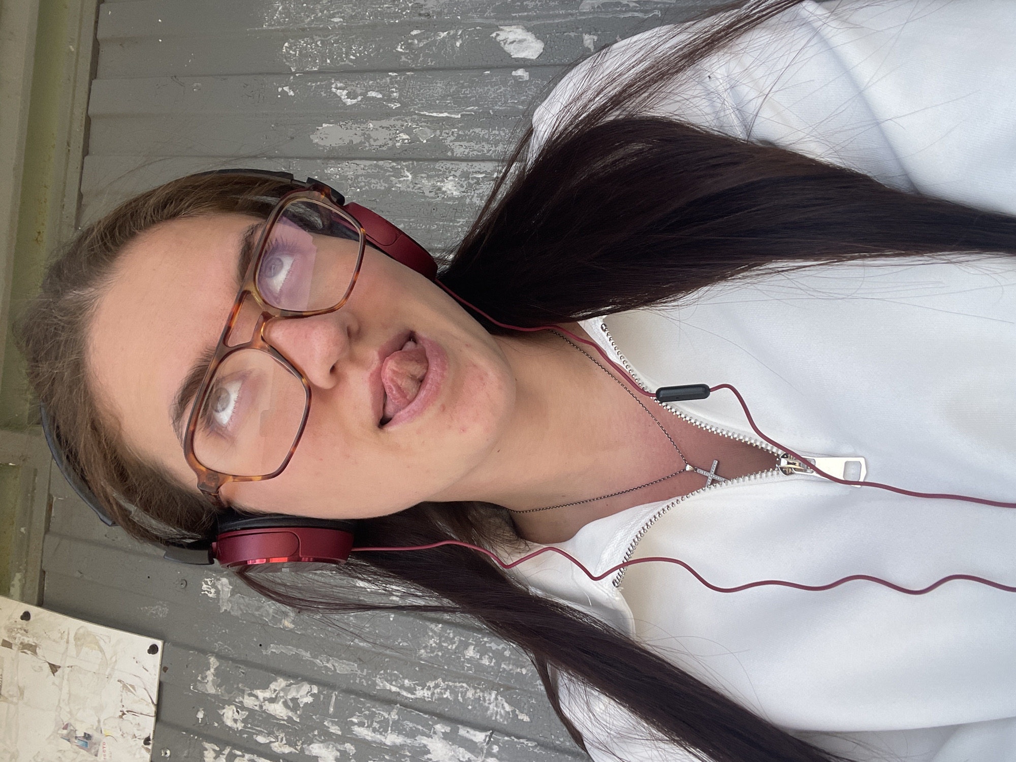 A young woman with long, dark brown hair is facing the camera, her head tilted to the left. She has fair skin, brown eyes, and is wearing tortoise-shell framed glasses. She is sticking her tongue out slightly. Red headphones are around her neck, connected to wires that lead to her white, zippered jacket. A silver necklace with a cross pendant is visible. The background appears to be a weathered, gray surface with peeling paint, possibly an exterior wall. The location is Molokovo, Russia.