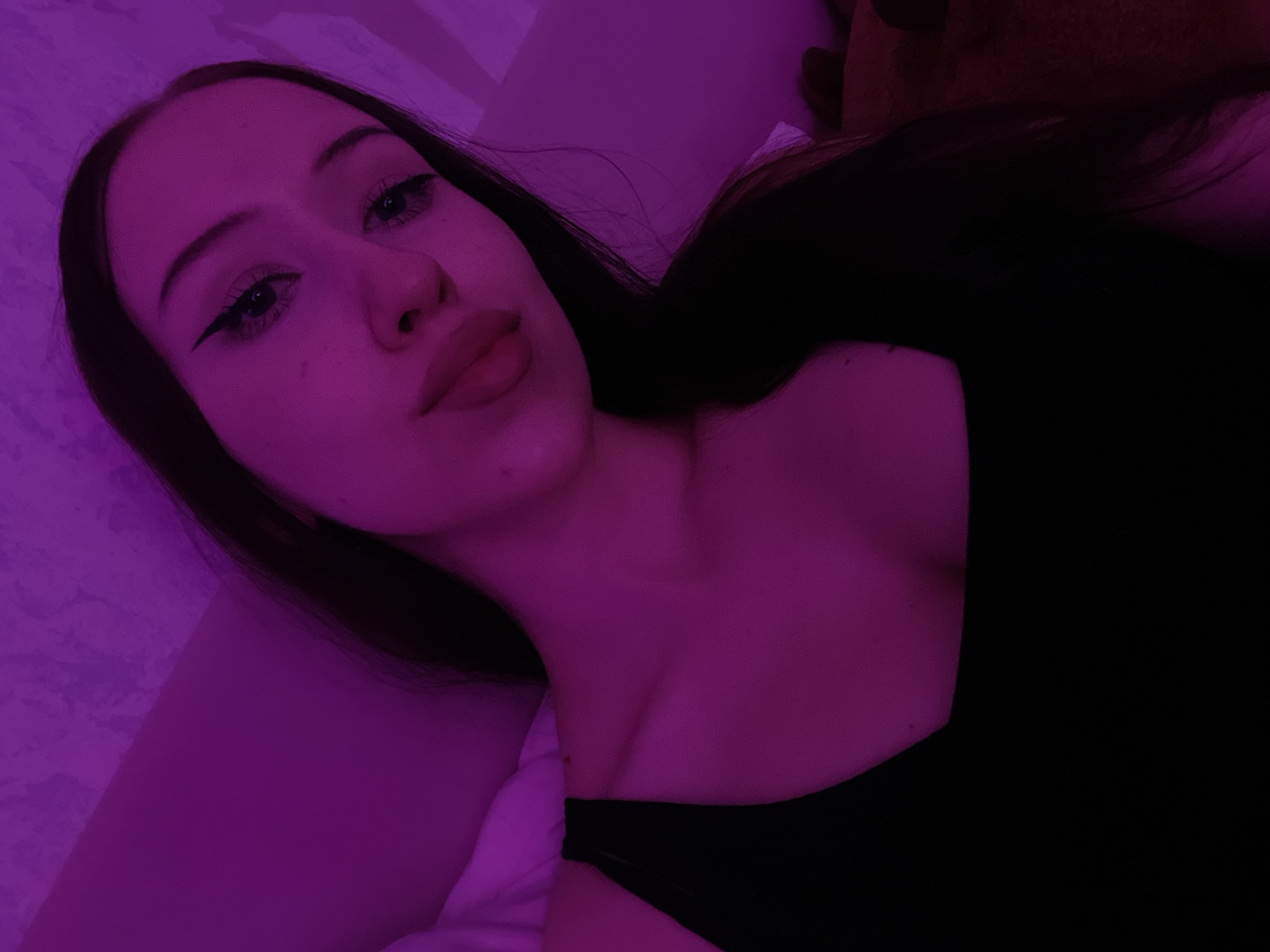 A young woman with long dark hair and eye makeup is taking a selfie in a room with purple lighting. She is wearing a black tank top and her head is tilted slightly. Her facial expression is neutral. The background appears to be a wall and possibly a bed. There are no discernible landmarks or indications of the specific location within Votkinsk, Russia.