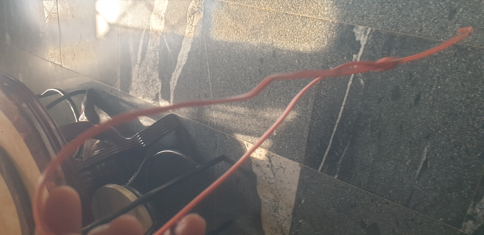The image shows a close-up of a person's hand holding a red rubber band. The rubber band is twisted and knotted. The background is a tiled floor with a dark gray and white marble pattern. Sunlight is casting shadows across the floor. In the upper left corner, a portion of a wooden table and chair legs are visible. The scene appears to be indoors, possibly in a home or public space.