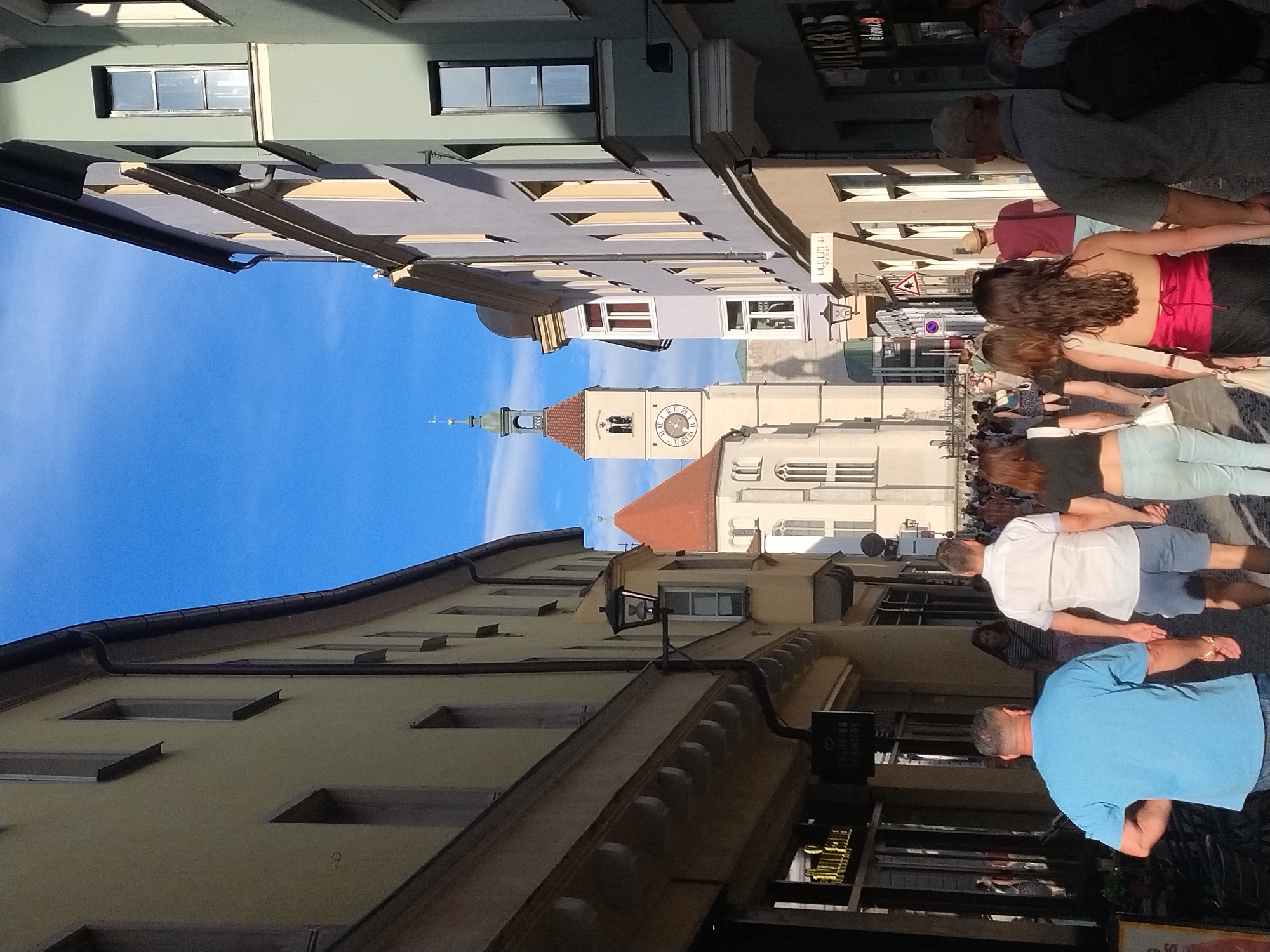 The image depicts a view down a narrow, pedestrianized street in Regensburg, Germany, lined with colorful, historical buildings. The architecture suggests a European city center, with light-colored stucco facades, pitched roofs, and numerous windows. A prominent clock tower, likely a landmark such as the Old Town Hall tower (Altes Rathaus), stands in the background, its clock face visible. Sunlight casts shadows on the buildings, indicating it is daytime, and the sky is a clear blue.
Several people are walking in both directions along the street, some in small groups and others individually. They are dressed in casual summer attire. A man in a bright blue t-shirt and jeans is walking away from the camera, and a couple holding hands, a woman in a black top and light blue jeans and a man in a white shirt and blue shorts, are walking in the same direction. Another couple, a man in a maroon t-shirt and a woman in a red top and black pants, are walking towards the camera. Their interactions are not discernible beyond their proximity and movement.
Shops with awnings and displays are visible on either side of the street, suggesting commercial activity. Some signs for businesses are partially visible. The street is paved with cobblestones, typical of older European city centers. The overall atmosphere is one of a busy, lively public space on a pleasant day.