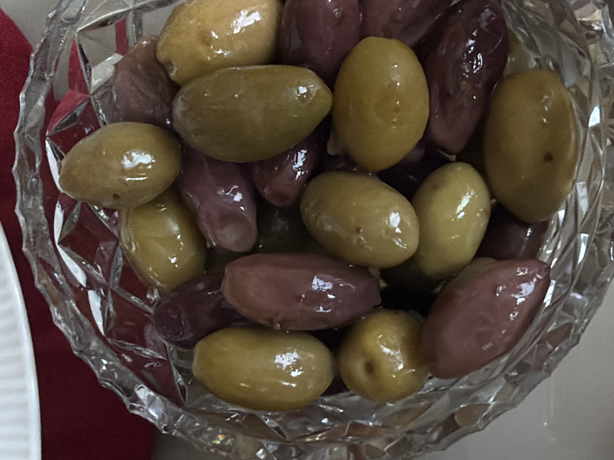 A close-up, high-angle shot depicts a clear, cut-glass bowl filled with a mixture of olives, primarily green and purple varieties, appearing shiny with a light coating of oil or brine. The bowl is situated on a surface with a red fabric visible on the left and a white surface peeking in from the lower left corner. The olives are arranged densely within the bowl. There is no indication of a location in Putilkovo, Russia.