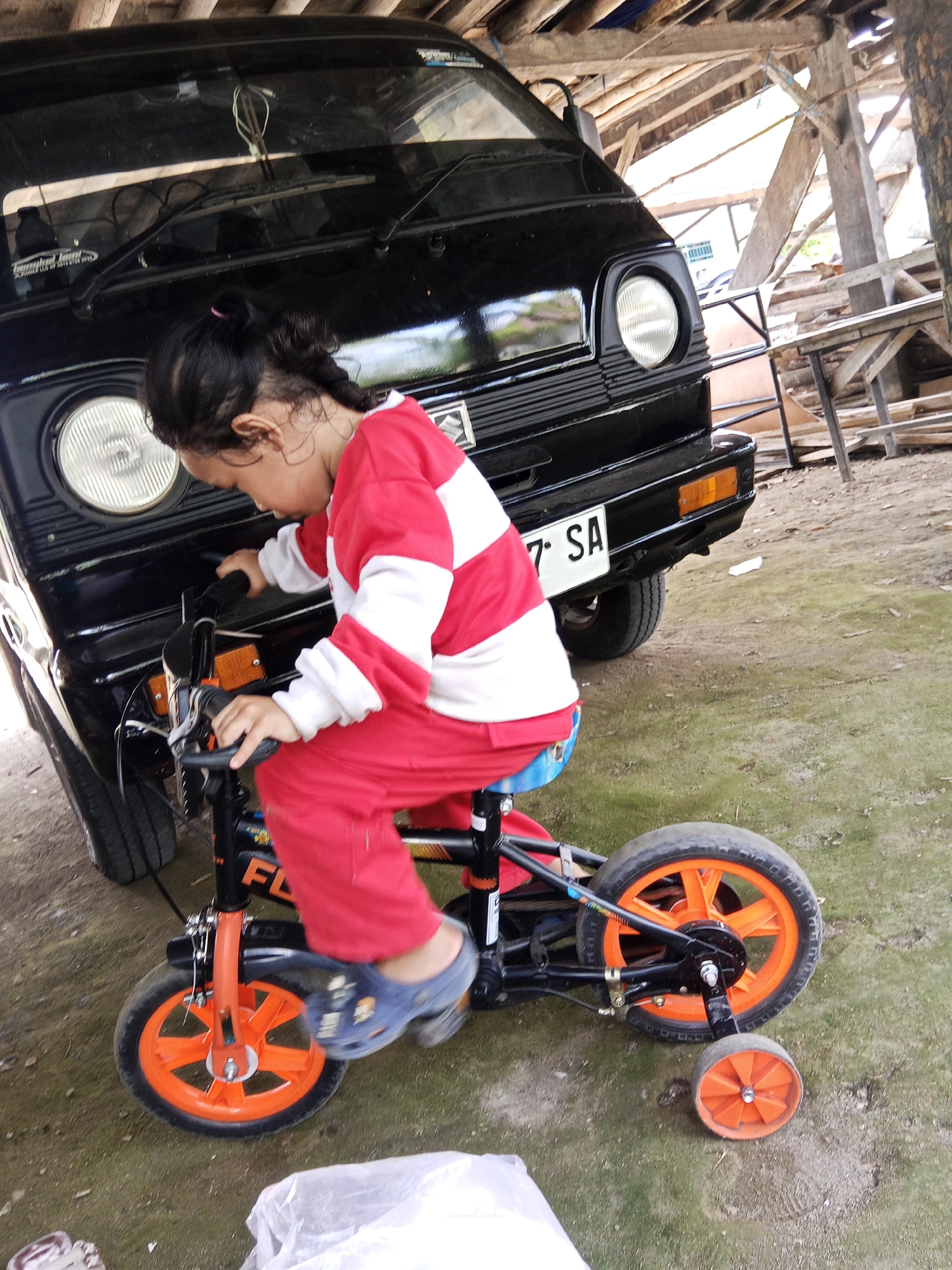 A young child, appearing to be female, is sitting on a bicycle in front of a black vehicle. The child is wearing a red and white striped long-sleeved shirt and red pants. They are looking down at the handlebars of the bicycle. The bicycle is orange and black and has training wheels. The black vehicle appears to be a van or a small truck. The scene is outdoors on a dirt and grassy ground. In the background, there is a wooden structure, possibly a shed or a part of a building, with exposed beams. There is also evidence of other wooden materials and debris in the background. The lighting suggests it is daytime. The license plate on the black vehicle is partially visible and reads "7 SA". This scene is located in a rural or semi-rural area, consistent with the outskirts of Lubuklinggau, Indonesia, where such outdoor settings with vehicles and simple structures are common.