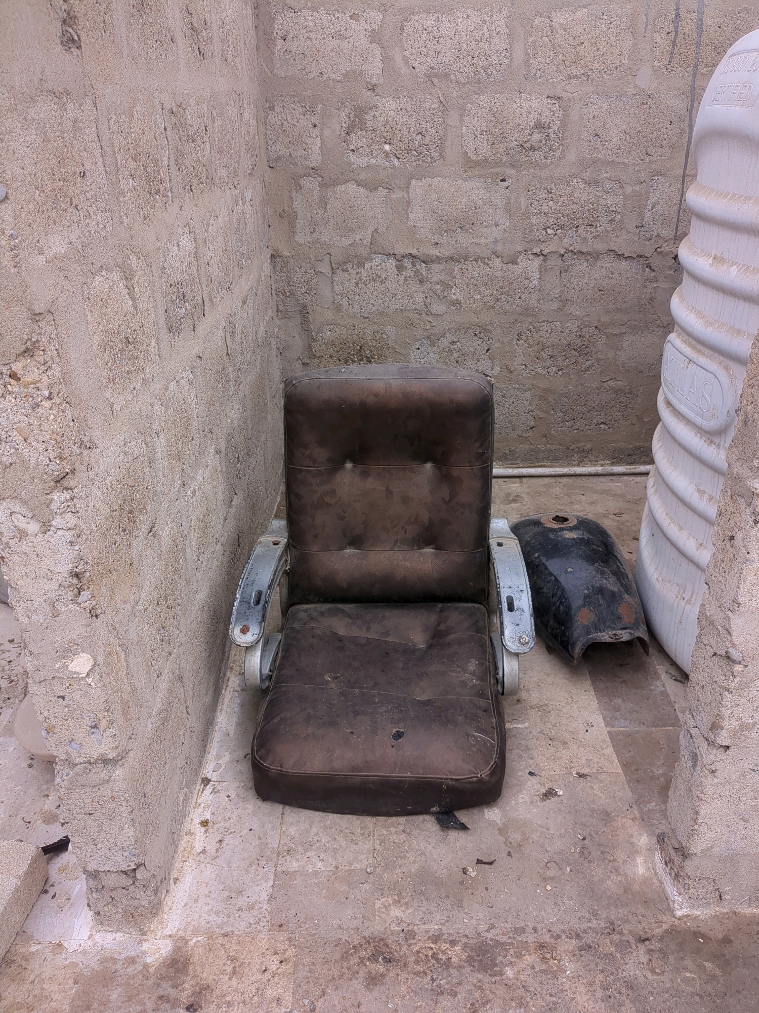 An outdoor scene shows an old, worn brown leather armchair with metal armrests situated on a tiled floor. Next to the armchair is a black motorcycle fuel tank, also appearing weathered. The background consists of a rough, textured concrete wall. To the right of the armchair is a large, ridged white plastic tank. The setting suggests an urban environment, possibly an alleyway or storage area, within the city of Karachi Division, Pakistan.