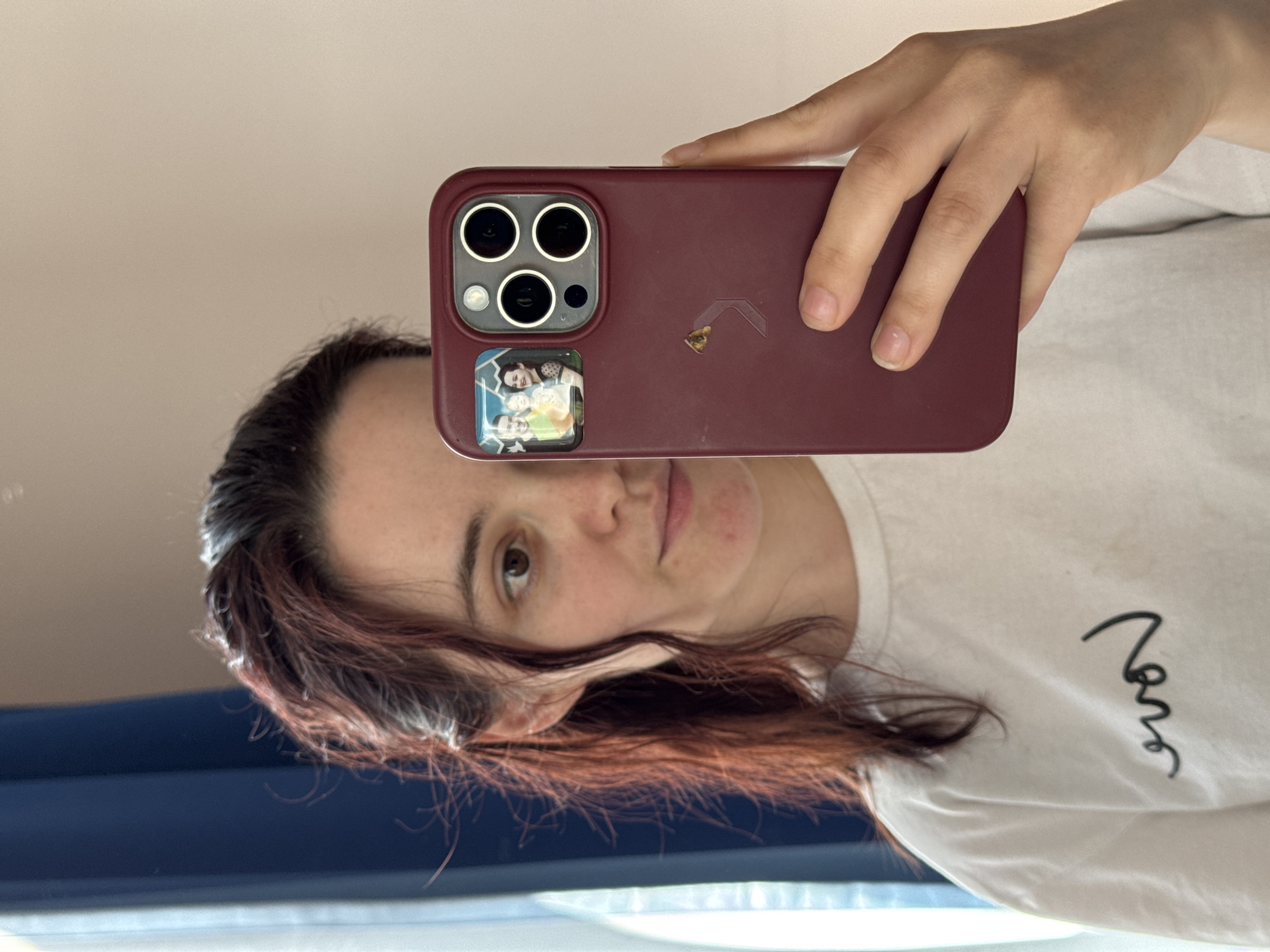 A woman is taking a selfie in a mirror using a maroon-colored iPhone. Her face is partially obscured by the phone. The phone has a sticker with a picture on its case and several camera lenses. She has dark hair with reddish highlights and is wearing a white t-shirt. The background appears to be a window with a blue curtain or blind. The location in the city of Sochi, Russia, is not discernible from the image.