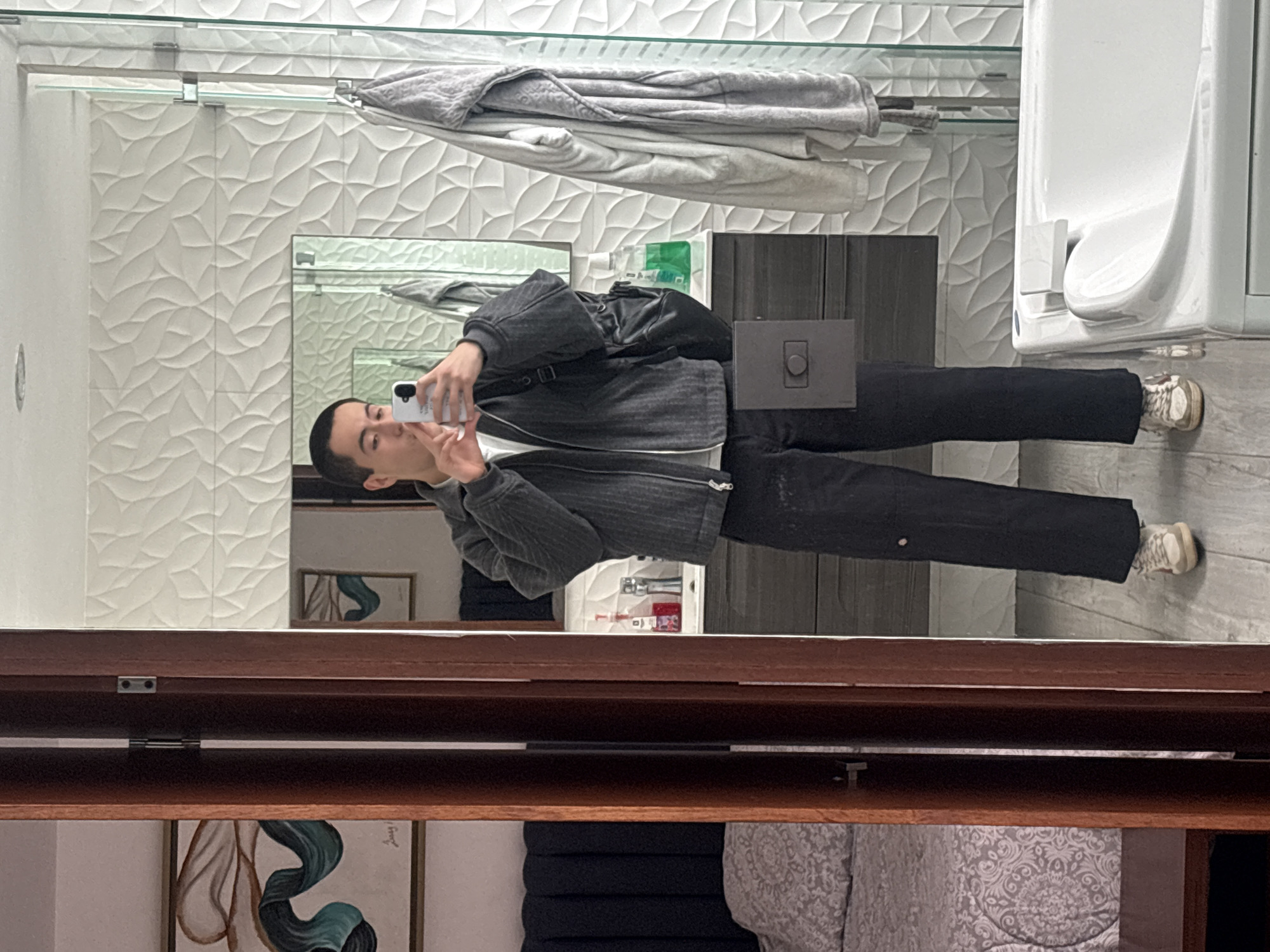 A person is taking a selfie in a bathroom. The person is wearing a dark jacket over a white t-shirt and black pants. They have a black backpack on their right shoulder. The bathroom has white tiled walls with a leaf pattern, a dark vanity with a sink, and a white bathtub. A towel is hanging on a rack next to the bathtub. To the left of the frame, a portion of a bedroom is visible, showing a bed with a patterned comforter and a dark headboard. The overall scene suggests a modern, minimalist aesthetic.