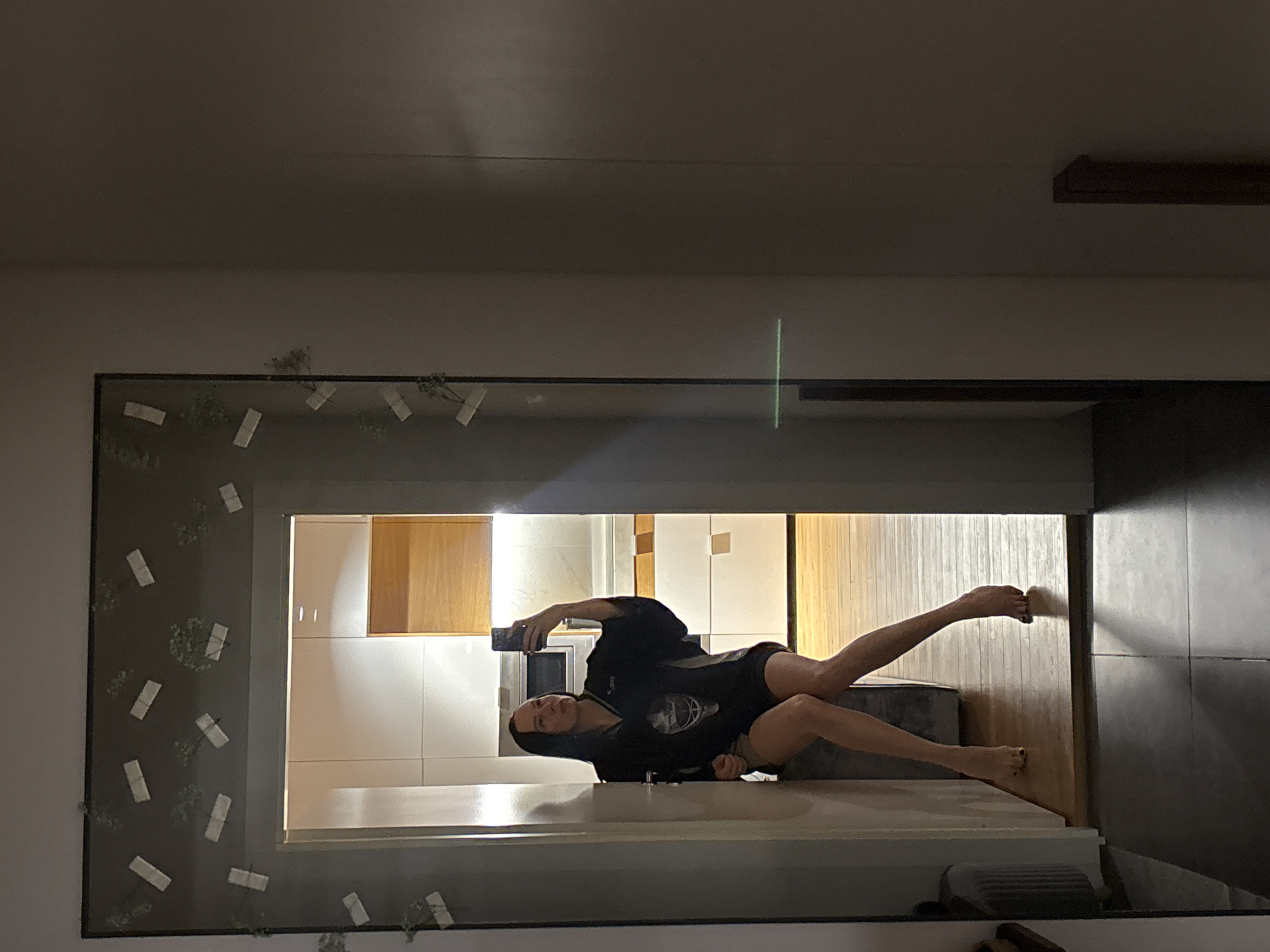 An indoor mirror selfie captures a person, likely a woman, lying in an unconventional pose within a modern residential setting in Chaikovsky, Russia. The individual is dressed in a dark hoodie or long-sleeved top and shorts, holding a smartphone to capture their reflection. They are positioned on a light-colored surface, possibly a kitchen counter or low platform, with their bare legs extended.
The background visible through the mirror's reflection shows a contemporary interior, characterized by white tiled walls and warm-toned wooden cabinets or panels, suggesting a kitchen or an open-plan living space. The mirror itself features a dark frame, which is decoratively adorned with several small, light-colored rectangular objects, appearing to be tape securing small green plant sprigs. A faint, vertical green light streak is visible in the upper part of the reflection, likely an optical artifact. The lighting suggests an indoor environment illuminated by artificial sources. There is no visible text in the image.