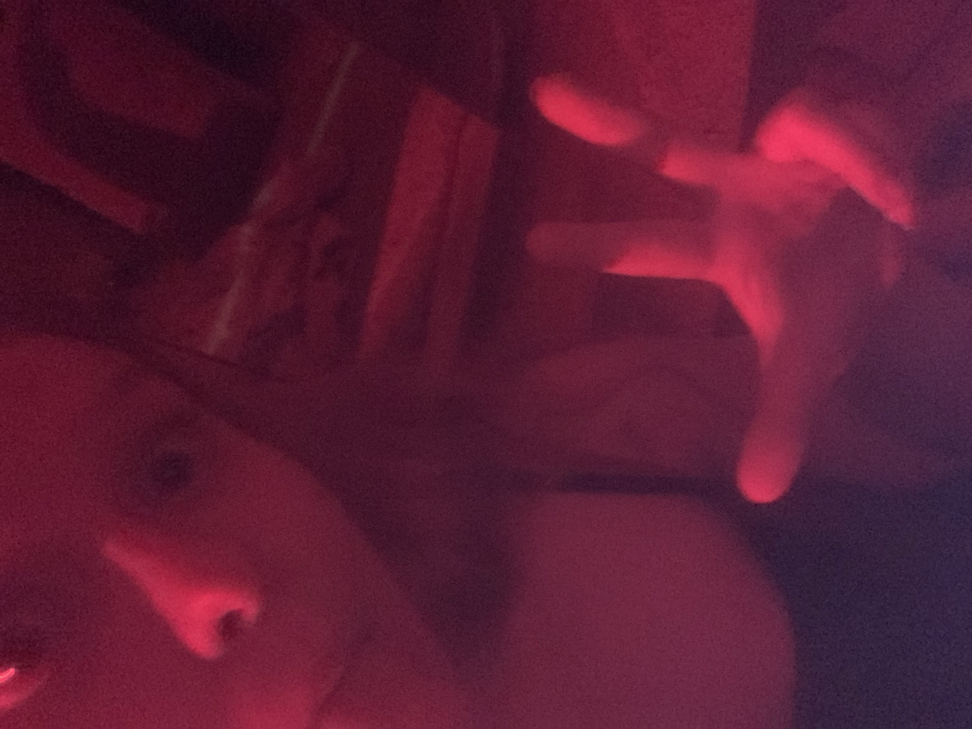 This dimly lit, soft-focus image is heavily cast in red light, creating a moody and intimate atmosphere in what appears to be an indoor setting. In the lower-left quadrant, a person's face is partially visible, appearing to look towards the viewer. Only a portion of the left side of the face, including the nose and parts of an eye and cheek, is discernible, softly illuminated by the red light. From the upper-right side of the frame, a hand is extended outwards, with fingers spread, brightly highlighted by the intense red illumination. The background consists of indistinct, dark shapes, suggesting an indoor environment with minimal ambient light other than the red source. The activity seems to be an interaction with the camera or a posed shot, with the hand reaching out in a gesture. There is no visible text. The scene implies a nighttime indoor environment, possibly a room with artistic or mood lighting, located in Vsevolozhsk, Russia.