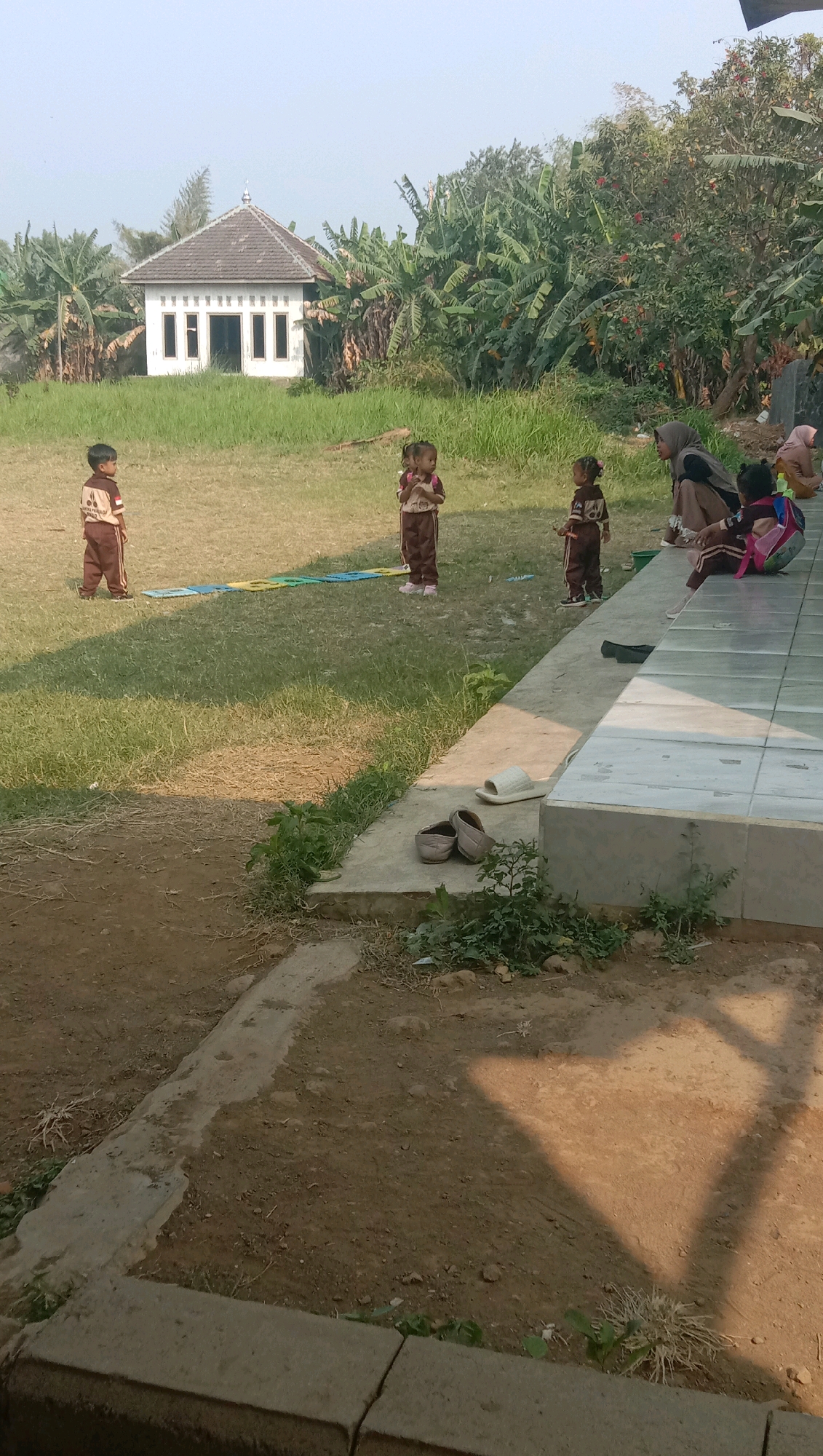 This image depicts an outdoor scene at what appears to be a school or community area in Karang Anyar, Indonesia. In the background, a small, white building with a gray, gabled roof stands amidst lush greenery, including banana trees. In the foreground, several children in school uniforms are scattered across a grassy field. To the right, a group of children and at least one adult are seated on a tiled patio. A few pairs of shoes and a single slipper are visible on the ground near the patio. The ground in the foreground is a mixture of dirt and some concrete elements, with shadows suggesting it is daytime and the sun is at an angle.