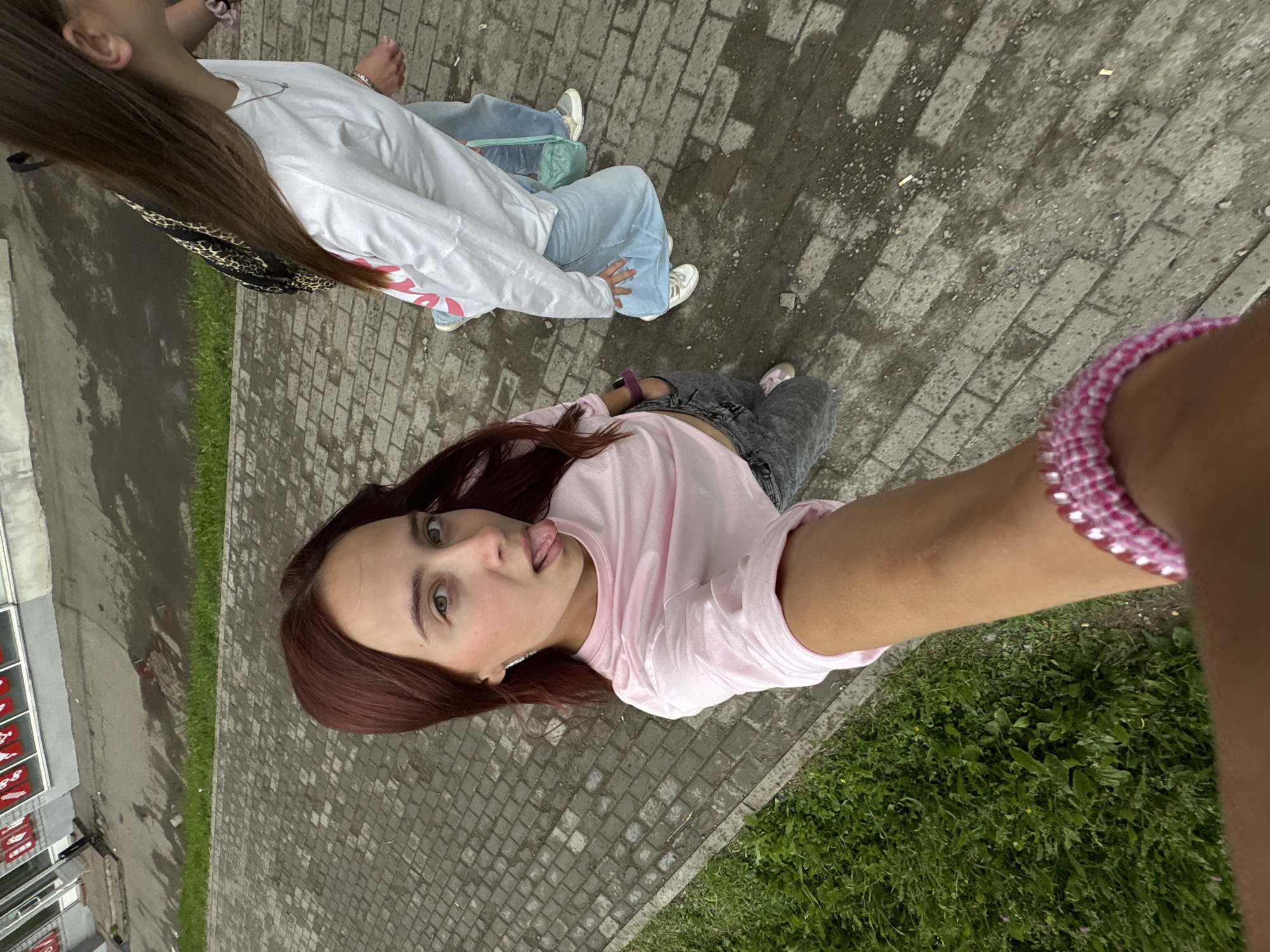 Two young women are walking on a wet cobblestone sidewalk in front of a building with red-trimmed windows. One woman, with long brown hair, is wearing a white long-sleeved shirt and light blue jeans. She has a leopard-print backpack slung over her shoulder and is wearing white sneakers. The other woman, with long reddish-brown hair, is closer to the camera and is taking a selfie. She is wearing a light pink cropped t-shirt, grey jeans, and pink sneakers. She has a pink beaded bracelet on her left wrist and is sticking out her tongue. The environment appears to be urban, with a grassy area to the left of the sidewalk. The presence of a building with retail displays and the cobblestone street suggests this is likely a commercial or pedestrian area within the city of Izhevsk, Russia, though specific landmarks are not identifiable.