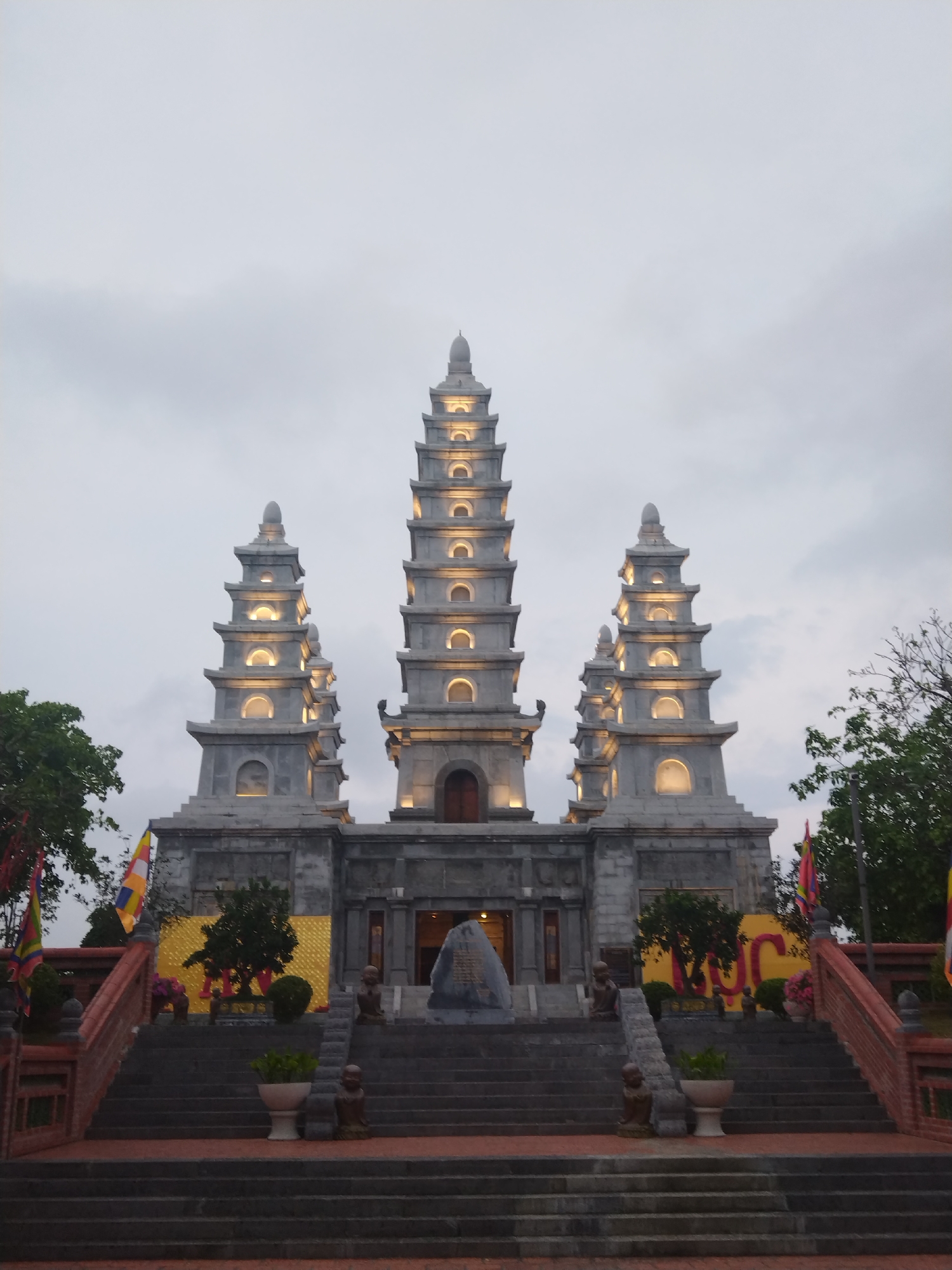 The image shows a Buddhist temple in Hong Gai Ward, Vietnam. The temple complex consists of three pagodas, the central one taller than the two flanking structures. The pagodas have a grey stone appearance, with tiers and arched openings illuminated by internal lights. The temple sits atop a series of ascending steps with brick-red railings, adorned with statues of monks. Flags and banners flank the staircase. A cloudy sky forms the backdrop.