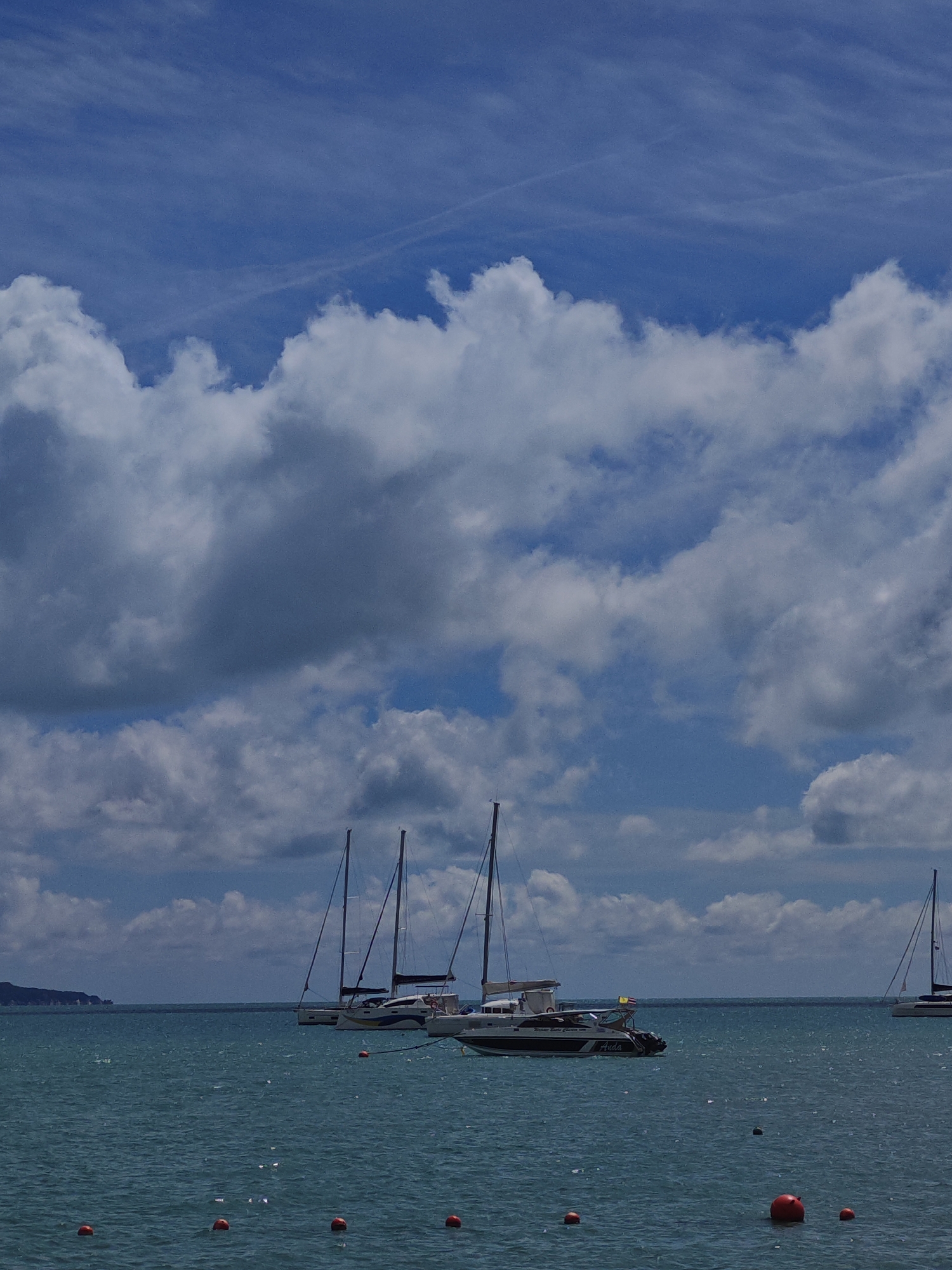 The image captures a serene scene of boats docked on the turquoise waters of Baan Bo Phut, Thailand. The sky is a vibrant blue, adorned with fluffy white clouds. Several sailboats are anchored, their masts reaching towards the sky, while a motorboat with the name "Auda" visible on its side is also present. The water is calm, with a few orange buoys floating on the surface, marking a designated area. In the distance, a lush green island adds to the picturesque backdrop. The overall atmosphere is one of tranquility and natural beauty.