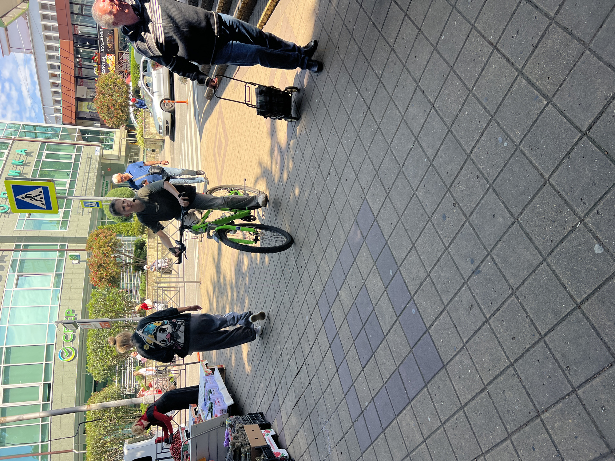 This image captures a vibrant street scene, likely in Sochi, Russia, under bright daytime sunlight. The ground is paved with interlocking grey and dark grey square tiles.
In the foreground, an older man with grey hair, wearing a dark, striped sweater and blue jeans, is seen pulling a black, wheeled trolley on the right side of the pavement. His silhouette casts a long shadow. Towards the center, a man in a dark t-shirt and grey pants stands beside a bright green bicycle, holding its handlebars. Further back on the sidewalk, another man in a blue shirt and jeans is standing.
On the left side of the frame, a street vendor's stall is set up, laden with crates of fresh produce, including what appears to be cherries and strawberries, with a box label showing "Клубника" (Strawberries). A person with blonde hair tied in a bun, wearing a black graphic long-sleeved shirt and grey sweatpants, stands near the stall, looking down. Another individual in a red top is bending over the displayed produce.
In the background, modern multi-story buildings with extensive glass facades line the street. One building clearly displays the Cyrillic logo "СБЕР" (Sber), indicating a major Russian bank. Other Cyrillic text, including "КУБАНИ," is visible on another building's facade. Trees and bushes add greenery to the urban landscape. A yellow triangular pedestrian crossing sign with a black symbol is visible on the left. Several other people can be seen walking or sitting on benches in the distance, and a light-colored car is on the road behind the individuals.
The scene depicts a typical daily hustle in a city environment, with people going about their routines of commuting and shopping under clear, sunny weather, as evidenced by the distinct shadows cast.
