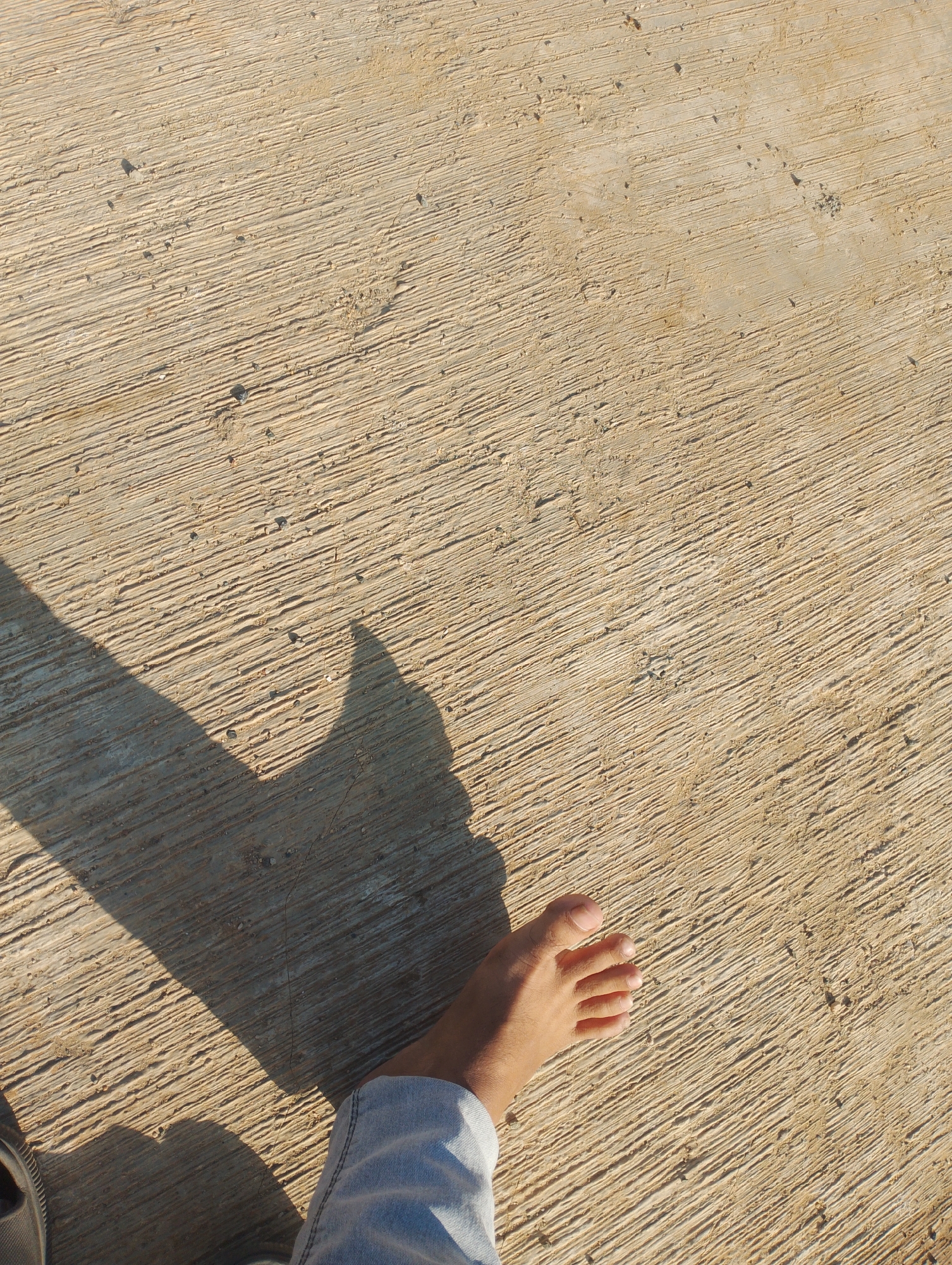 A person's left foot, clad in light blue denim jeans, is positioned on a textured, light brown concrete surface. The concrete exhibits parallel grooved lines, indicative of its construction or finishing. A shadow, cast from an unseen object to the left, stretches across the lower left portion of the frame, forming a distinct triangular shape. Another, smaller shadow, possibly from the person's head and torso, is visible above and to the right of the foot. The lighting suggests it is either early morning or late afternoon, as indicated by the long shadows and warm tones on the concrete. The scene appears to be outdoors, with the surface potentially being a walkway, patio, or roadside in Batulicin, Indonesia.