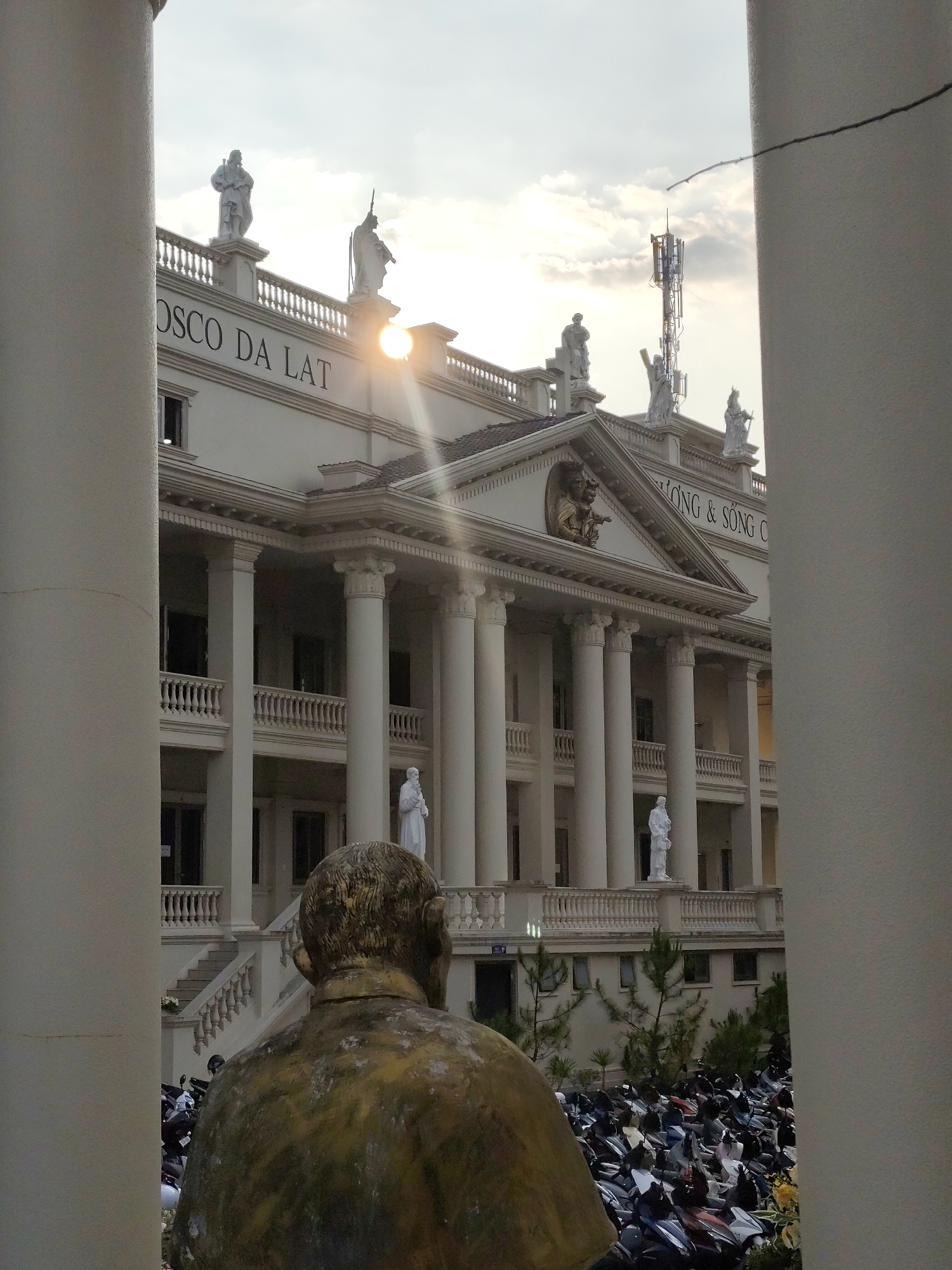 The image shows a partial view of the architecture of the Don Bosco Da Lat school in Da Lat, Vietnam, framed by two vertical white columns. The multi-story building has a classical style, featuring white columns and a facade with statues. The school’s name, "Don Bosco Da Lat," is written on the building's face. Statues adorn the rooftop and balconies. The sun is visible above the building, creating a lens flare.
A bronze bust is positioned in the foreground with its back to the viewer. Below the building, many parked motorcycles are visible. A telecommunications tower is near the building.