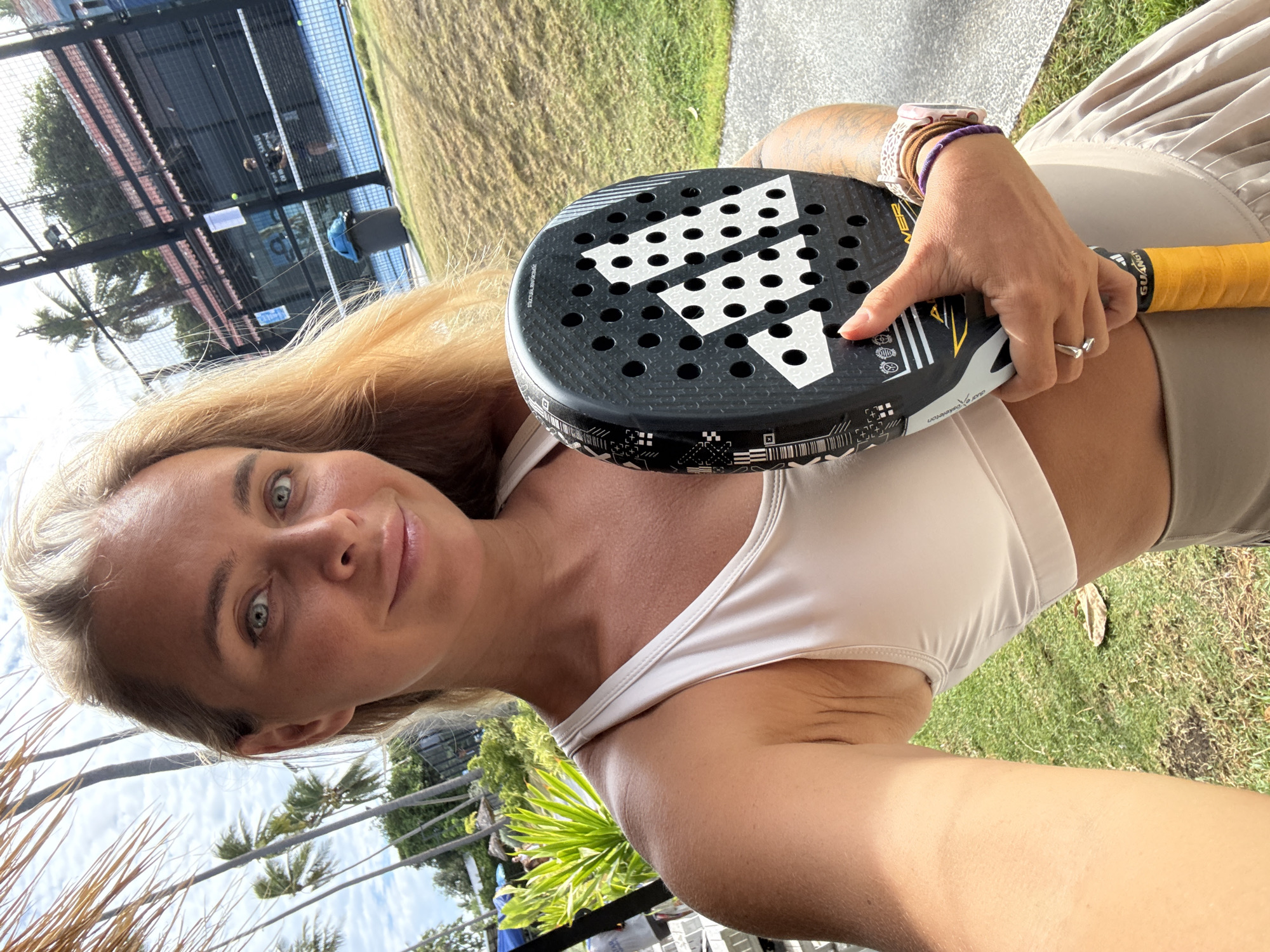 A woman with blonde hair takes a selfie while holding a black Adidas padel racket with a yellow grip. She is wearing a light beige sports bra and a beige pleated skirt. She has blue eyes and a slight smile. In the background, there is a blue padel court with people playing. The court is surrounded by a fence, palm trees, and lush green vegetation. The sky is partly cloudy. The scene appears to be a sports facility in a tropical location, likely Ko Samui District, Thailand, given the presence of palm trees and the sunny weather.