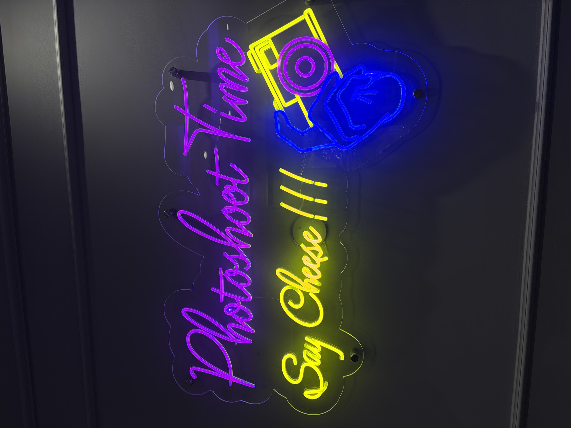 A neon sign with the words "Photoshoot Time" in purple and "Say Cheese !!!" in yellow is mounted on a dark gray wall. To the right of the text, a neon illustration depicts a blue hand holding a yellow camera with a purple lens and body. The sign is supported by clear acrylic backing and secured with metal screws. The environment appears to be an indoor space, likely a studio or a themed room, in Sweileh, Jordan.