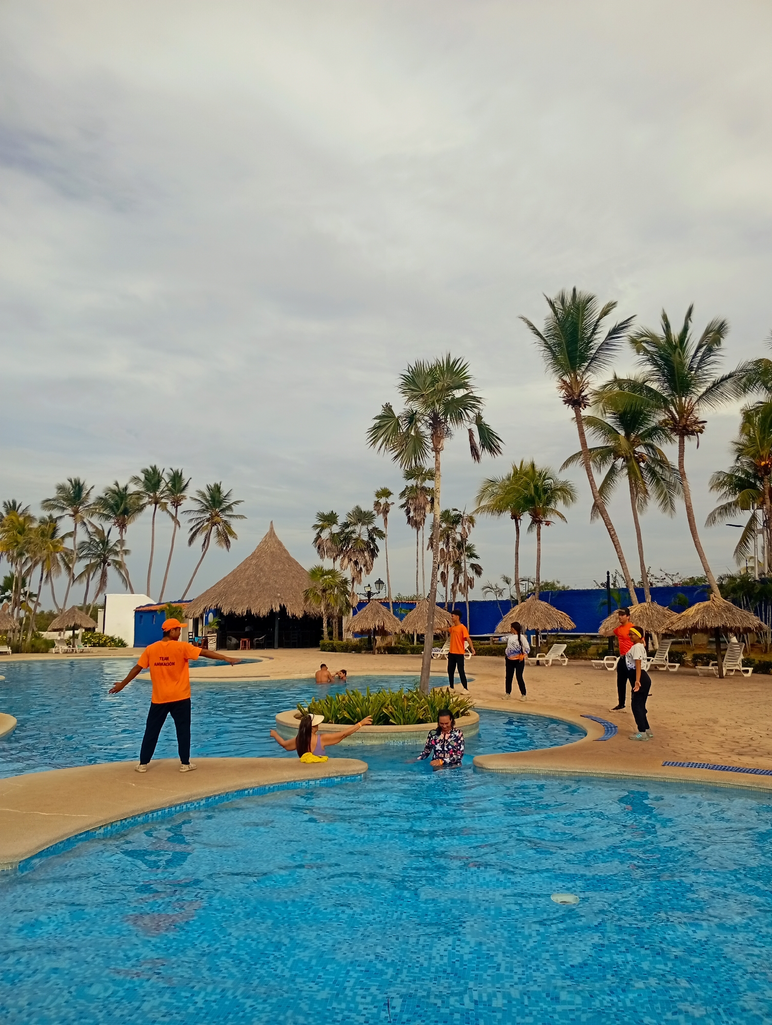 The image depicts a swimming pool area at a resort in Playa El Agua, Venezuela. The pool is segmented into multiple sections with concrete pathways connecting them. Several people are in the pool, and staff members wearing orange shirts are on the perimeter and pathways. There are palm trees, thatched-roof structures, and beach chairs. The sky is overcast. Some individuals are swimming and interacting within the pool area. Other individuals stand on the pool deck facing the pool.