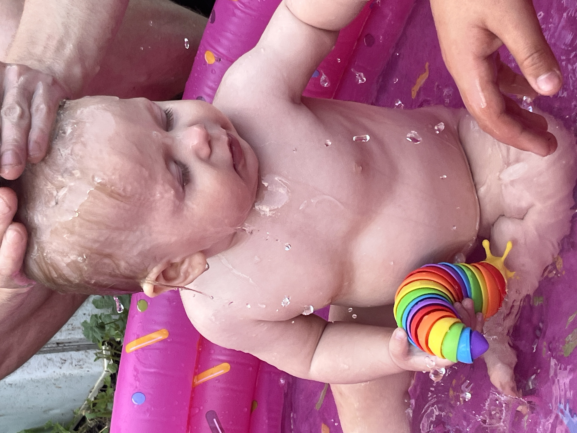 A baby, with eyes closed and looking down, is seated in a pink inflatable pool filled with purple water. The baby is wet, with water droplets visible on its skin and hair. A person's hands are visible above the baby's head, seemingly washing it. The baby is holding a colorful, segmented snail-shaped toy in its left hand. The pool has a donut-like pattern on its inflated rim, with colored circles and orange stripes. The background is blurred, showing some green foliage and a wooden fence. The scene is outdoors, suggesting a backyard or garden setting. There is no information about the location in the city of Фоки, Russia.