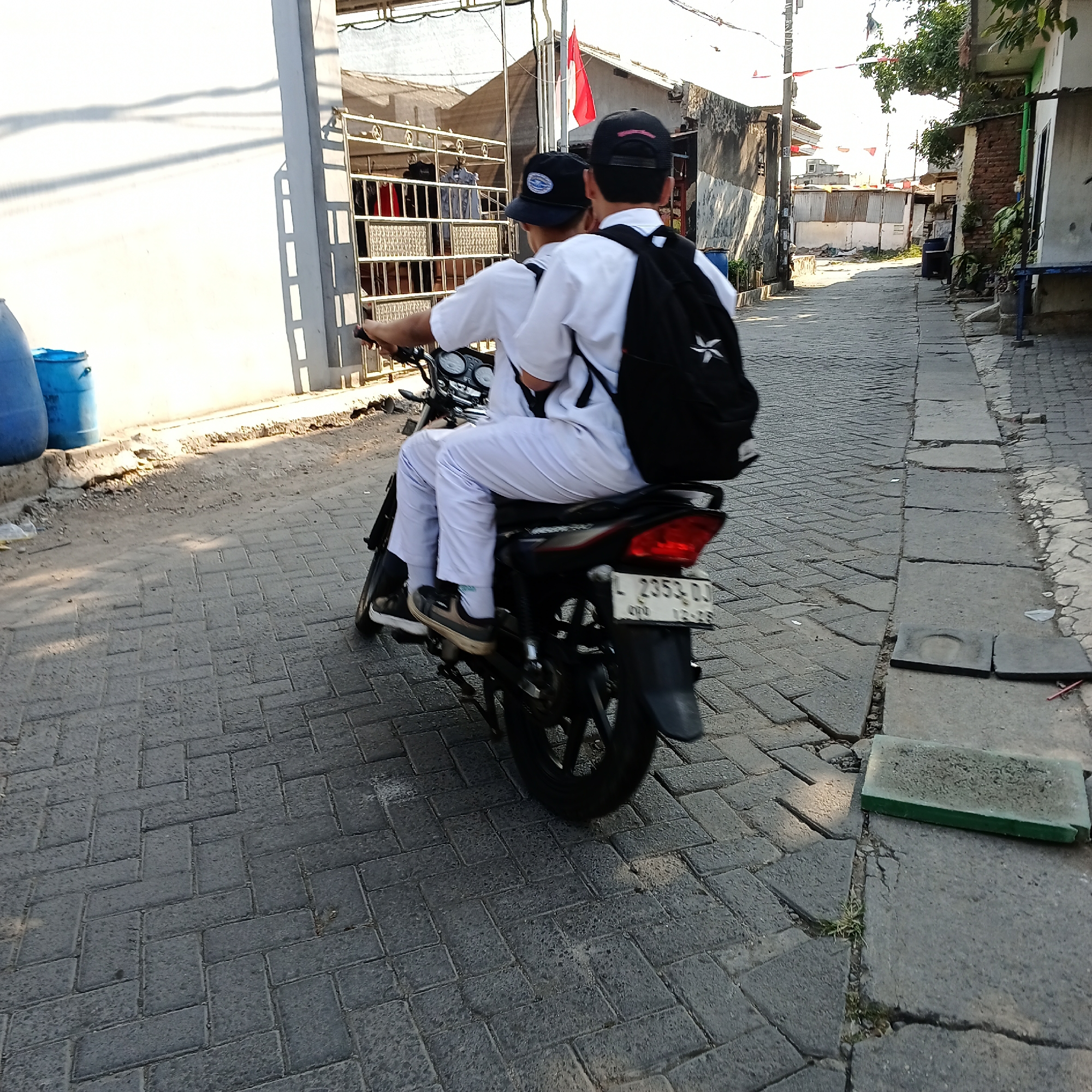 Two individuals wearing white uniforms and black backpacks are riding a motorcycle down a paved alleyway in Kepuh Kiriman, Indonesia. The motorcycle has a license plate that reads "L 2353 DD". The alley is bordered by buildings with white and brick walls, some featuring corrugated metal roofing. There are blue barrels visible on the left side of the image. The ground is paved with interlocking bricks, and a broken concrete drainage covers is on the right. The scene suggests a typical urban environment in the specified location.