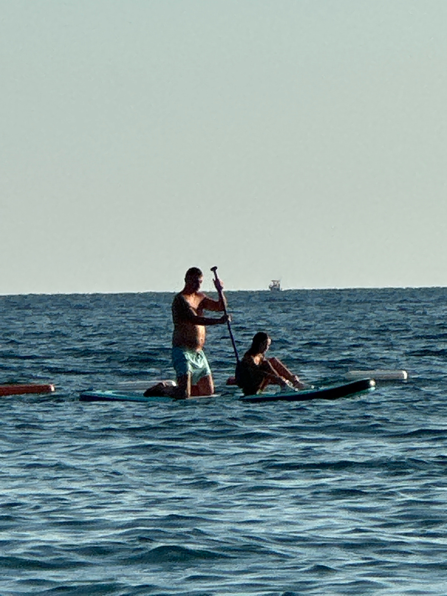 The image depicts a serene ocean scene with two individuals on paddleboards in the city of Boreti, Montenegro. In the foreground, a man is kneeling on a paddleboard, holding a paddle. To his right, a second person is seated on another paddleboard. The water is a deep blue, with visible ripples. In the distance, a hazy horizon separates the sea from a light-colored sky. A small boat is visible on the horizon. The overall impression is one of leisure and outdoor activity on the water.