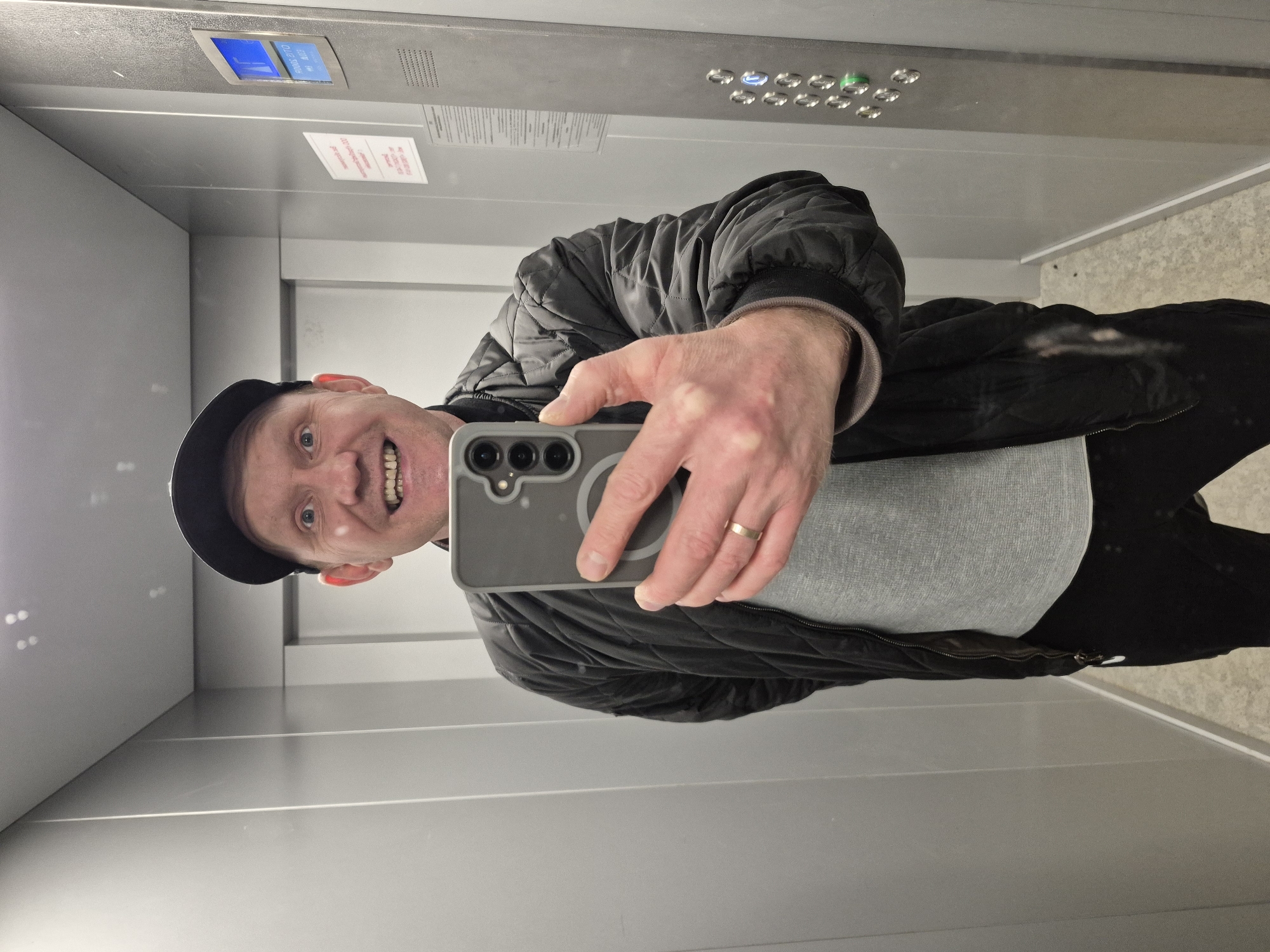 A man is taking a selfie in an elevator. He is wearing a black cap, a black quilted jacket over a gray t-shirt, and dark trousers. He is holding a gray smartphone with three camera lenses in his right hand. He is smiling and looking directly at the camera. He has a gold ring on his ring finger. The elevator has a gray metal interior with a digital floor display and control buttons. There is a small white information sticker on the wall. The location is presumed to be Chaikovsky, Russia.