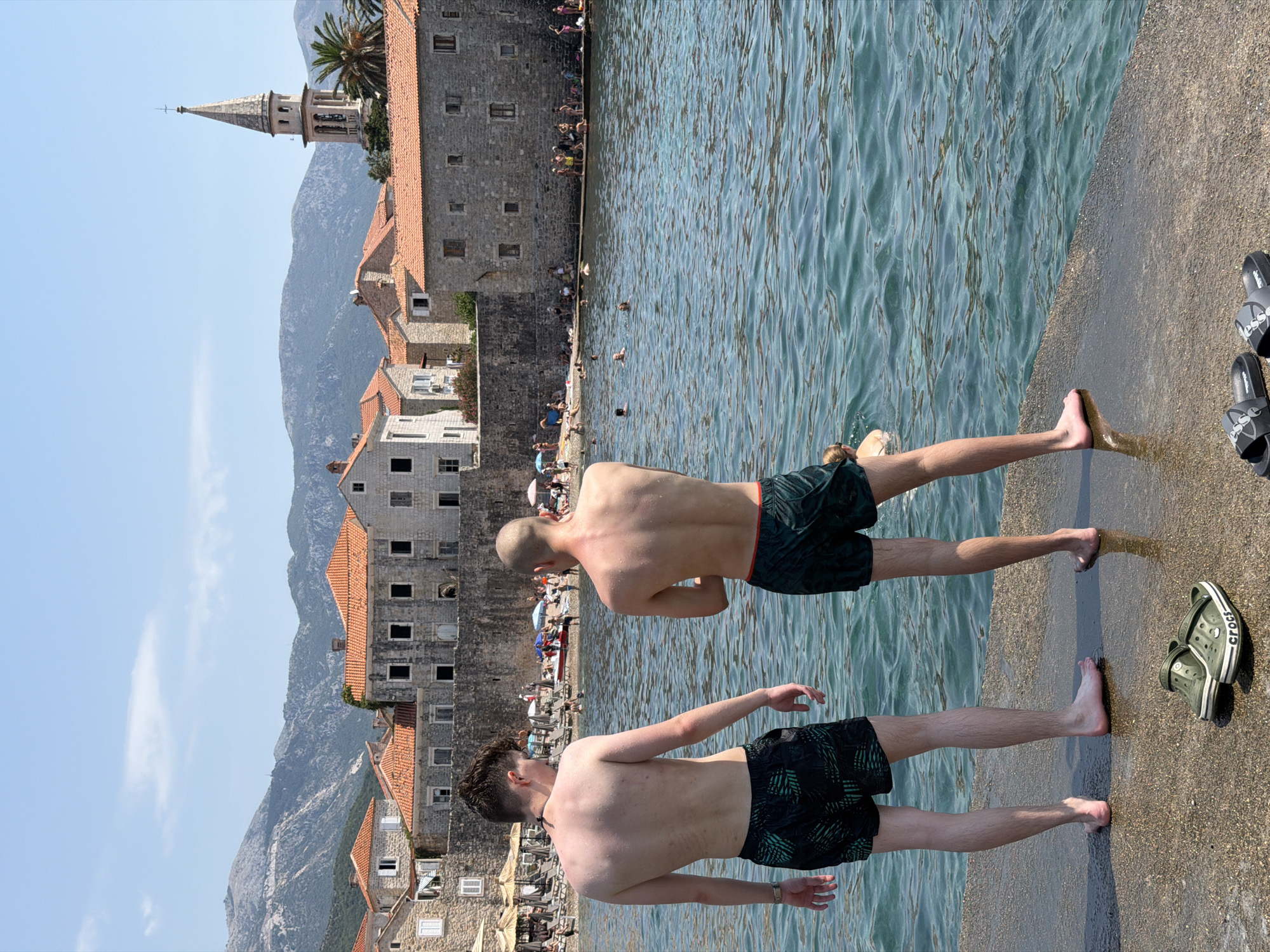 Two young men, viewed from behind, stand on a stone pier facing the turquoise water of the Adriatic Sea in Budva, Montenegro. Both are wearing dark patterned swim trunks. The man on the left has wavy brown hair and a watch on his left wrist. The man on the right has short, light-colored hair. In the foreground, a pair of green Crocs and a pair of black slide sandals lie on the wet pier. Beyond the water, the ancient walled Old Town of Budva is visible, with buildings featuring orange tile roofs. A bell tower rises above the cityscape, and mountains form the backdrop. Other beachgoers are visible in the distance, some in the water and others on the shore.