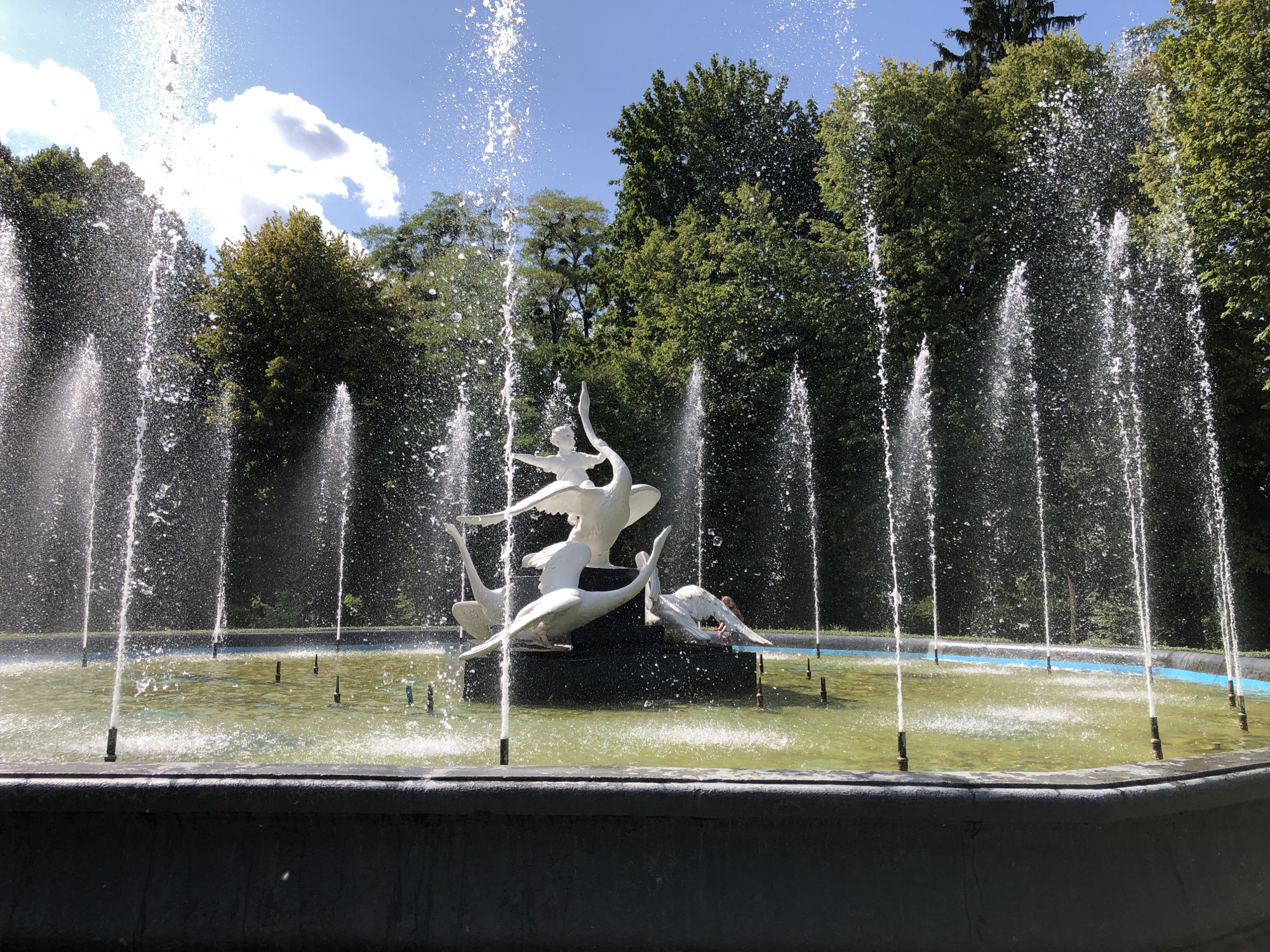 This image shows a fountain with a white sculpture of swans and figures in a park in Lviv, Ukraine. The fountain has multiple water jets spraying upwards, creating a misty effect. The sculpture appears to be a central feature of the fountain, depicting swans in various poses, some with human-like figures. In the background, lush green trees and a bright blue sky with white clouds are visible. The fountain is situated in a pool with greenish water. A person is partially visible in the background, near the fountain's sculpture. The fountain is located in Stryiskyi Park in Lviv.