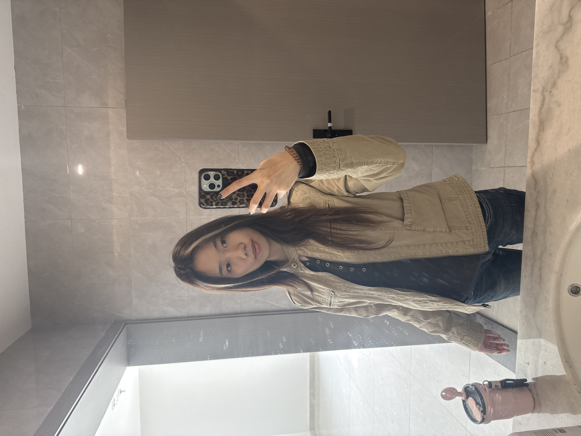 A person is taking a selfie in a bathroom. The bathroom has light grey tiled walls, a light brown cabinet, a white sink, and white walls. The person is holding a phone with a leopard print case and has long, dark hair. They are wearing a black shirt, jeans, and a tan jacket. Based on the background, the location is the Nan'an District of China.