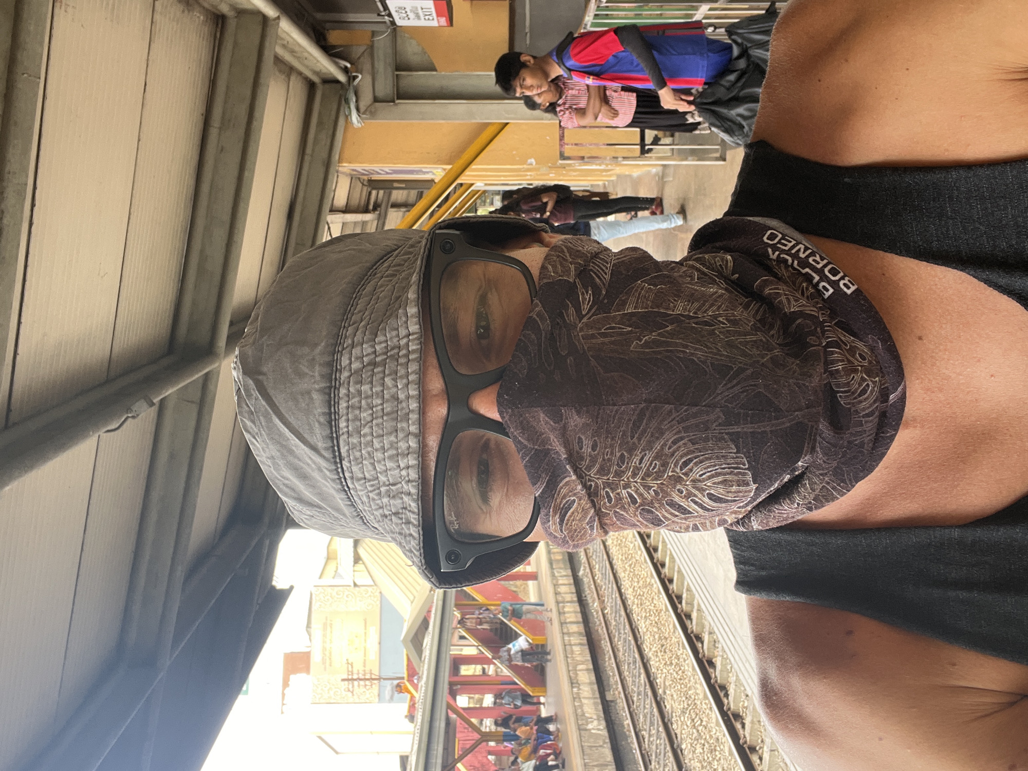 A first-person perspective selfie captures a person wearing a grey bucket hat, black-framed glasses, and a face covering with a brown leaf pattern at a train station. The individual is positioned in the foreground, looking directly at the camera. Behind them, a train track runs parallel to a covered platform where several people are visible. The platform structure is a mix of yellow and red metal supports and a corrugated metal roof. A sign with "EXIT" is visible on a yellow pillar to the right. The environment appears to be outdoors, with a bright, natural light source illuminating the scene. The backdrop suggests an urban setting with buildings visible in the distance.