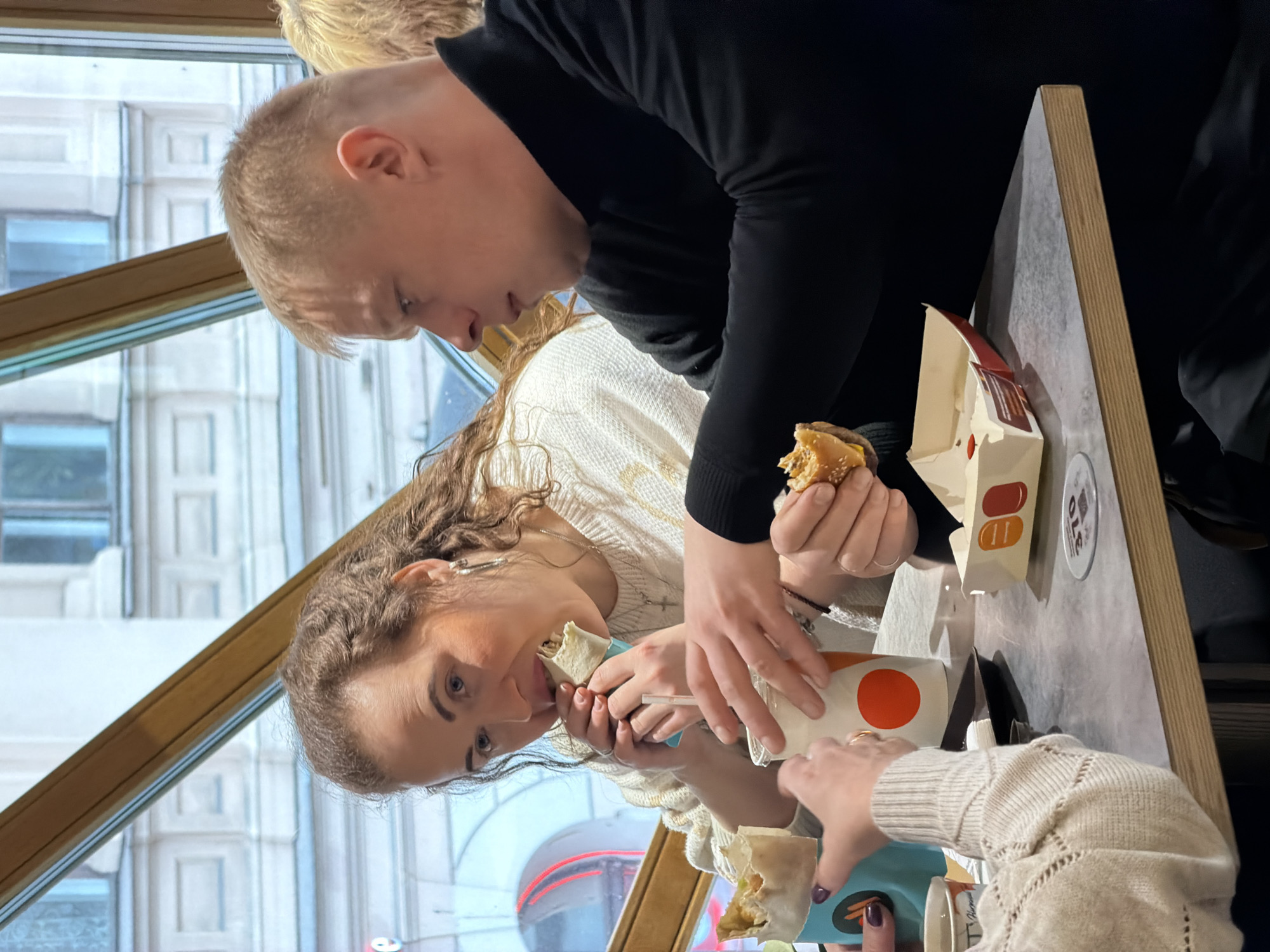 Inside a fast-food establishment in Saint Petersburg, Russia, three individuals are seated around a light grey table with a wooden edge. The location appears to be on an upper floor, offering a view through large windows of a classical-style building with multiple rectangular windows and stone architectural details. The establishment's branding, identified as "Vkusno i Tochka" by visible packaging, indicates it is a Russian fast-food chain.
In the center, a female with long, curly brown hair, wearing a cream-colored textured sweater, is actively eating a wrap, holding it with both hands to her open mouth. She wears silver hoop earrings and a silver cross pendant. To her right, a male with short, light brown hair, dressed in a black long-sleeved top, holds a partially consumed hamburger in his right hand, contained within a branded "Vkusno i Tochka" box. His gaze is directed towards his left, presumably at the female eating the wrap.
To the left of the central female, only the lower arms and hands of a third individual, also wearing a light-colored, possibly beige, cable-knit sweater, are visible. This individual holds a light blue cup containing another wrap and a white cup with an orange circular logo. The central female also holds a similar white cup with an orange logo, from which a straw protrudes.
On the table, a black tray holds napkins, and a circular coaster or table marker bearing the number "370" is visible. Outside the window, a reddish neon sign is faintly discernible in the distance. The building seen through the window is characteristic of the historical architecture prevalent in central Saint Petersburg, such as along Nevsky Prospekt.