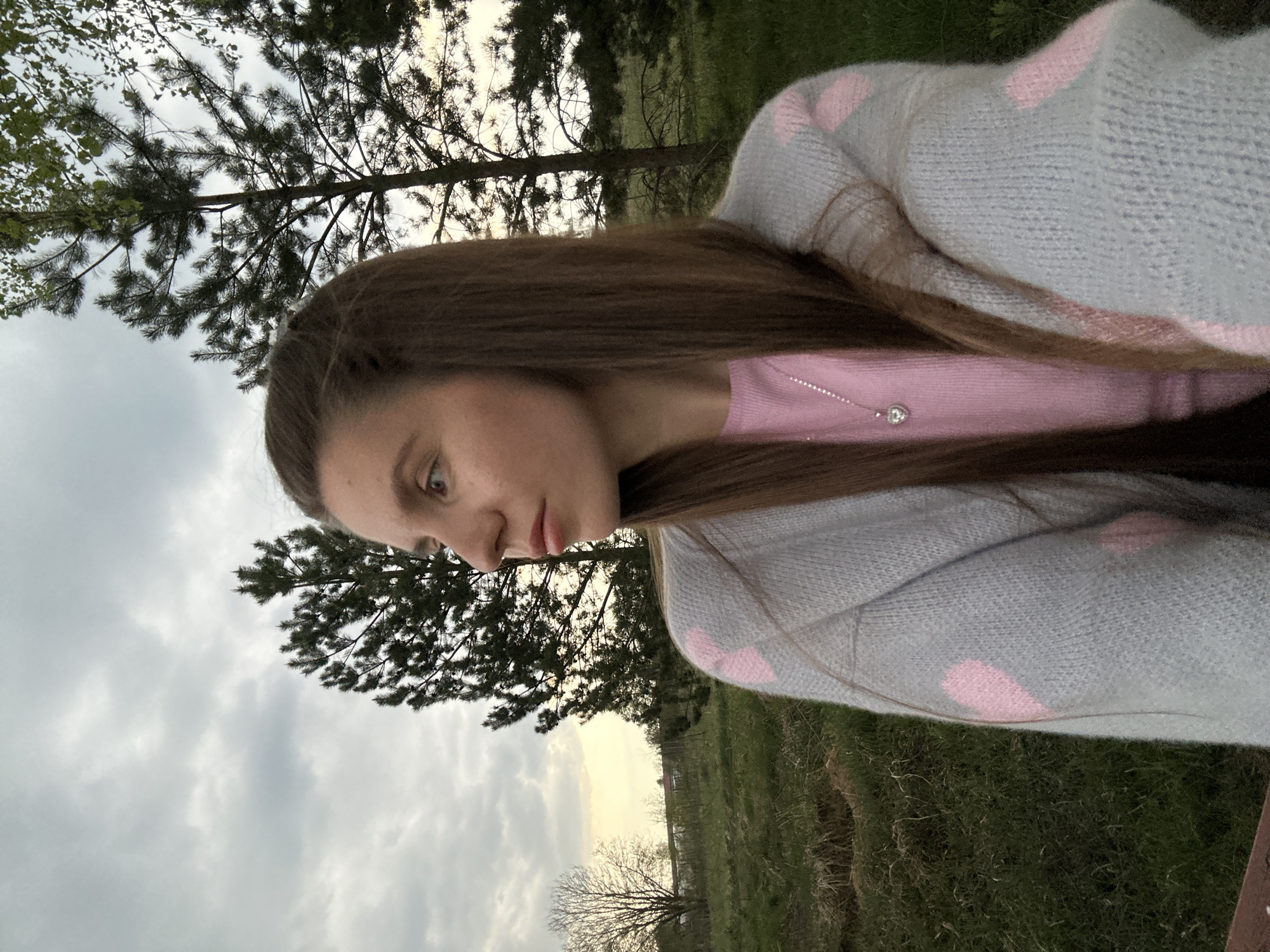 A young woman with long brown hair is outdoors. She is wearing a pink top and a light gray cardigan with pink heart patterns. The background consists of trees, a cloudy sky, and a grassy area. Given the prompt's constraint of location within Борщевка Малая, Russia, it's plausible the setting could be a rural or semi-rural area on the outskirts of the village.