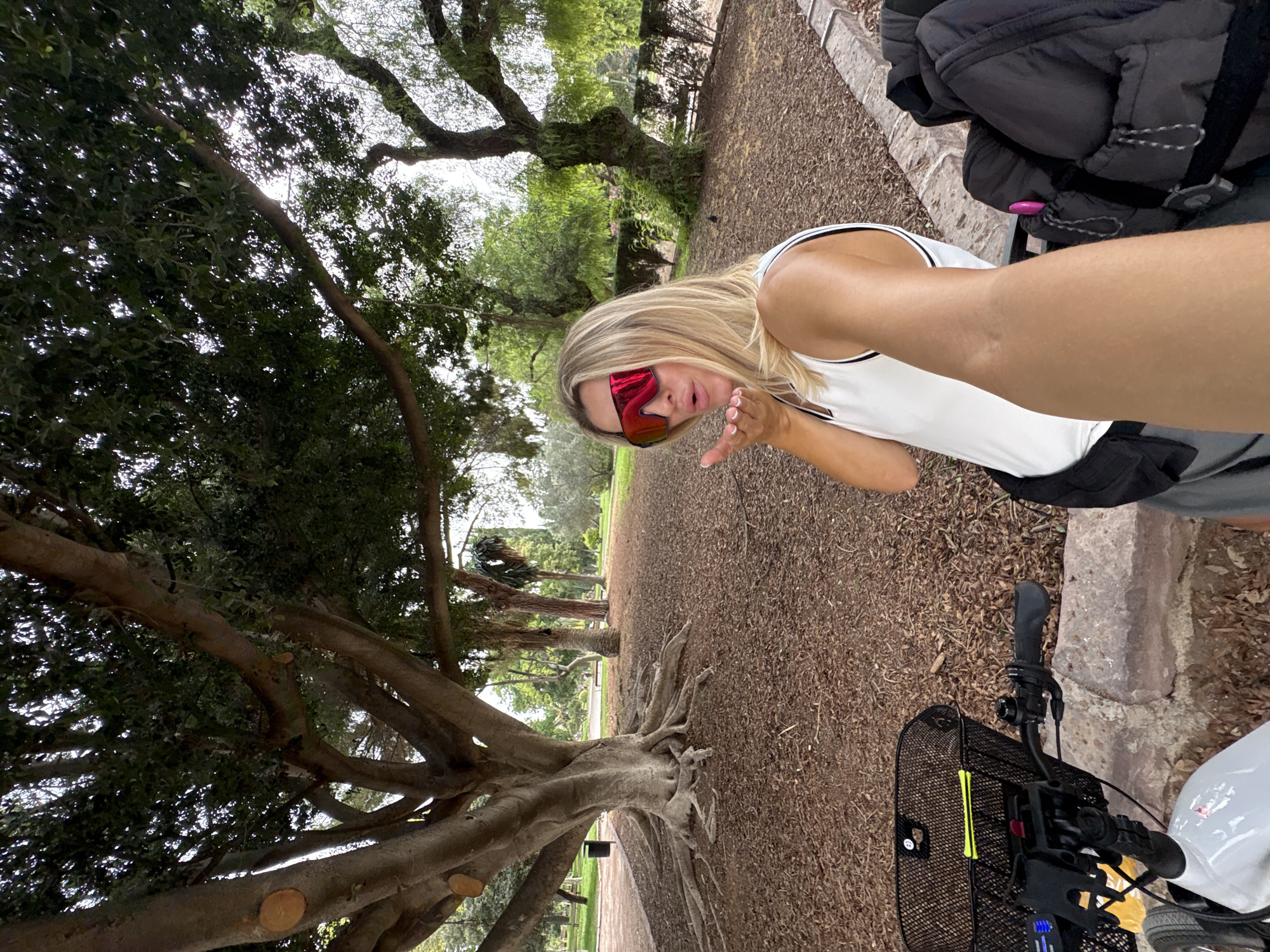 A blonde woman in a white athletic tank top and gray shorts wears large red sunglasses and blows a kiss towards the camera. She is in a park with a large tree with exposed roots to her left. The ground is covered in wood chips. To her right, a black backpack is visible, and a white electric scooter with a basket is in the foreground. The scene appears to be in Valencia, Spain, possibly the Turia Gardens, given the presence of mature trees and the general park-like environment.