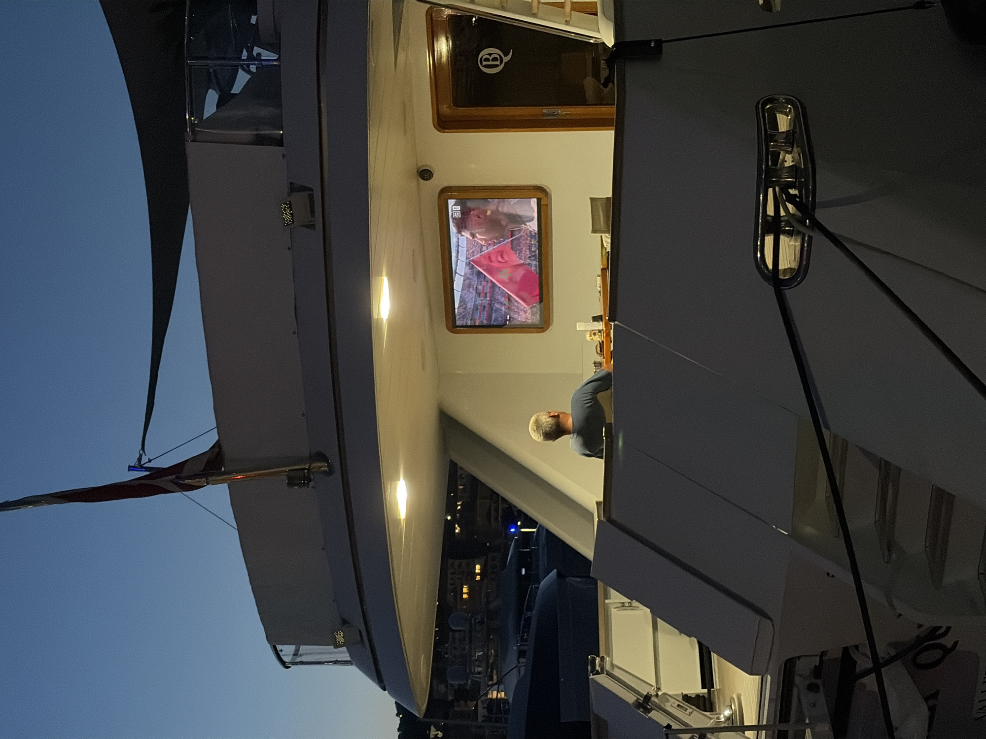 An exterior shot at dusk shows a person seated in front of a television on the deck of a yacht docked in Imperia, Italy. The person, a male with short gray hair, is wearing a blue shirt and is facing away from the camera, looking at the television. The television displays a sports broadcast, with a person holding a pink flag in the foreground. The yacht has a white hull and upper structure, with a dark-colored canopy overhead. In the background, other yachts are visible, as well as buildings with illuminated windows along the waterfront. The sky is a deep blue, indicating twilight. The overall scene suggests a relaxing evening on the water.
