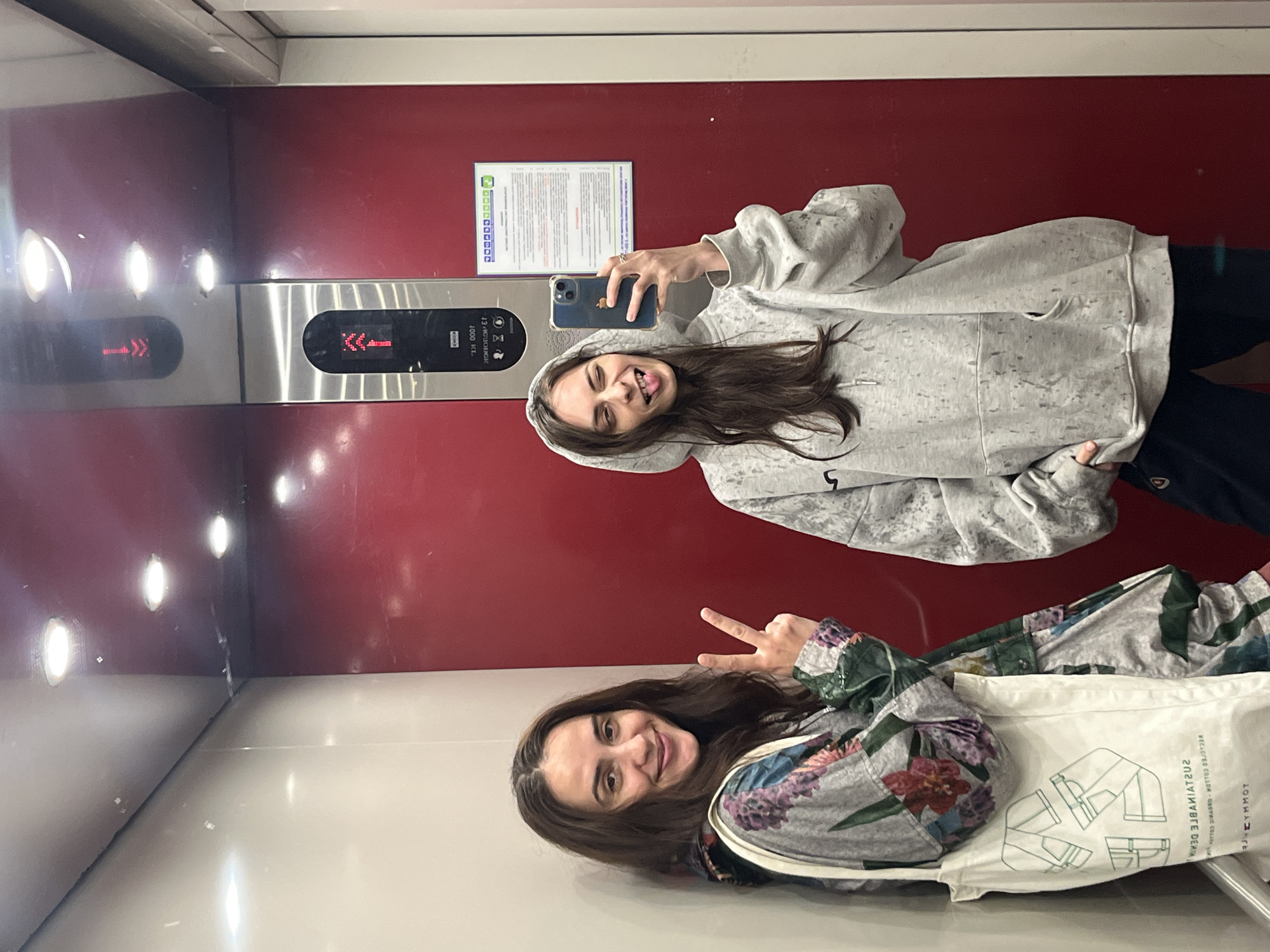 Two women are in an elevator. One woman is taking a selfie, holding a smartphone. She wears a light gray hoodie and black pants. The other woman sits on the floor, wearing a patterned floral shirt, making a peace sign. She has a white tote bag with sustainable denim information printed on it. The elevator has a red wall, a stainless steel control panel with an arrow display, and a white information sheet on the wall. The location is Krasnoyarsk, Russia based on the text in the elevator’s information sheet.