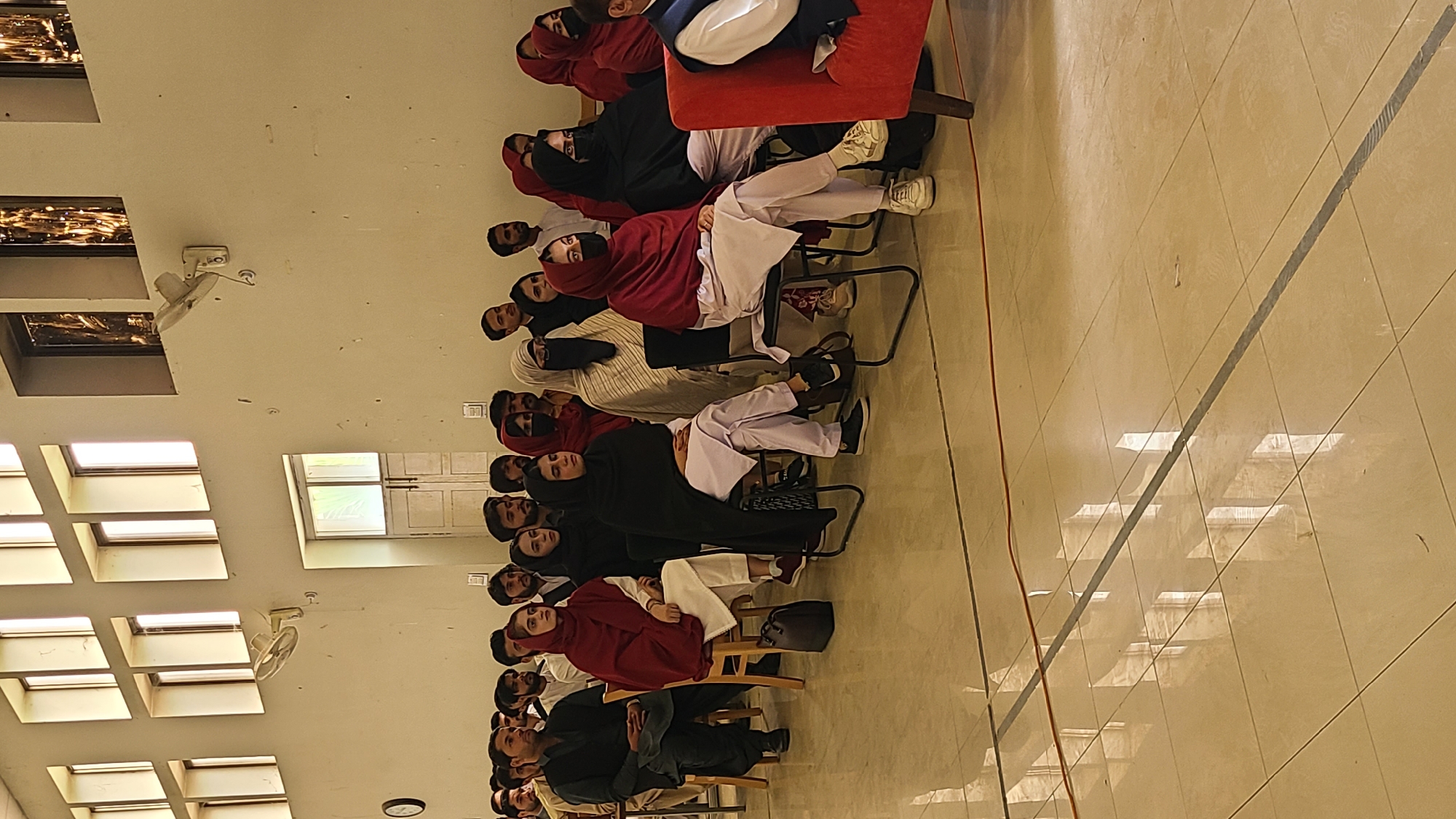 The image depicts a group of people, predominantly women wearing headscarves and some men, seated in rows of chairs in what appears to be an indoor event space. The women are wearing various colors, including red and black, and some wear white trousers. The men wear darker colored clothing. The setting is a large room with tile flooring and a high ceiling. Multiple windows are visible along one wall, and ceiling fans are mounted above. An armchair is visible at the far end of the row. The location is possibly in Quetta City Tehsil, Pakistan.